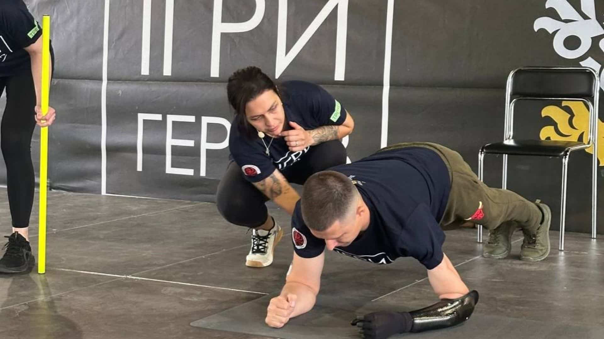 Військовий із протезом руки виконує вправу «планка», а інструкторка поруч підтримує його
