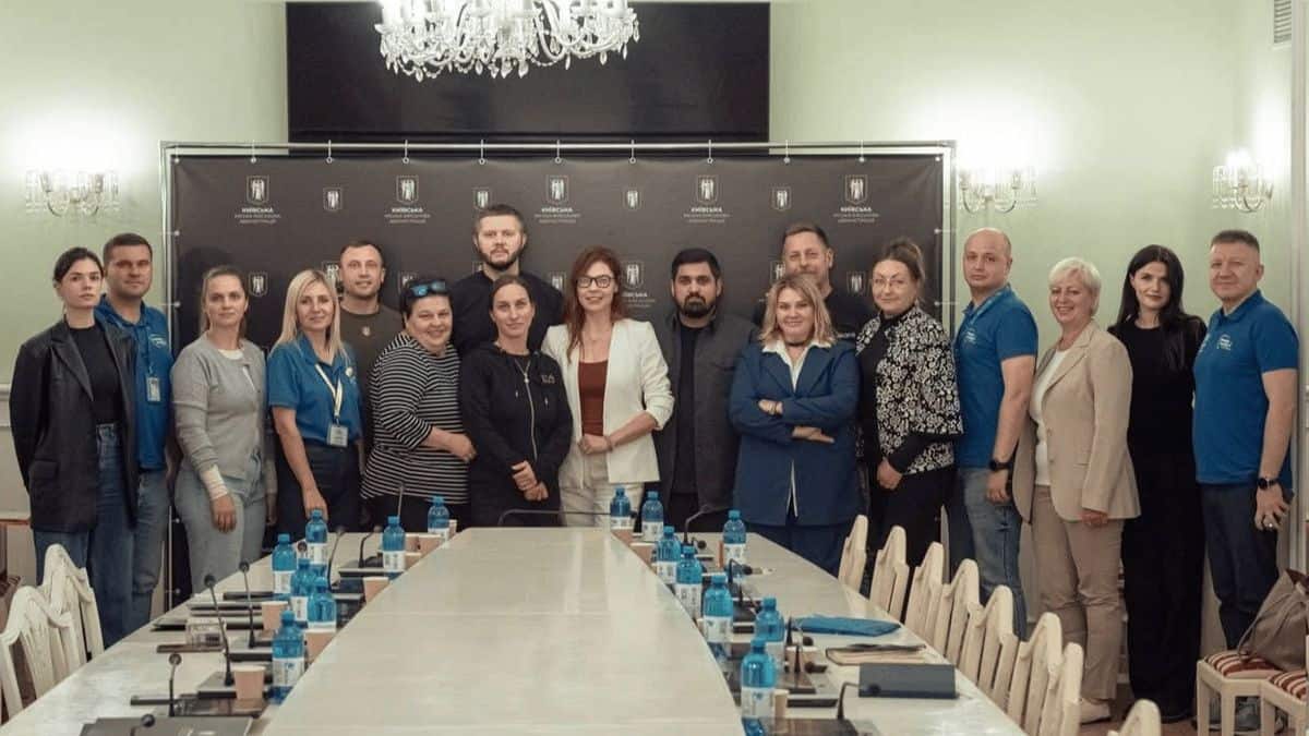 Група людей стоїть у залі для нарад перед довгим столом на фоні банера Київської міської адміністрації