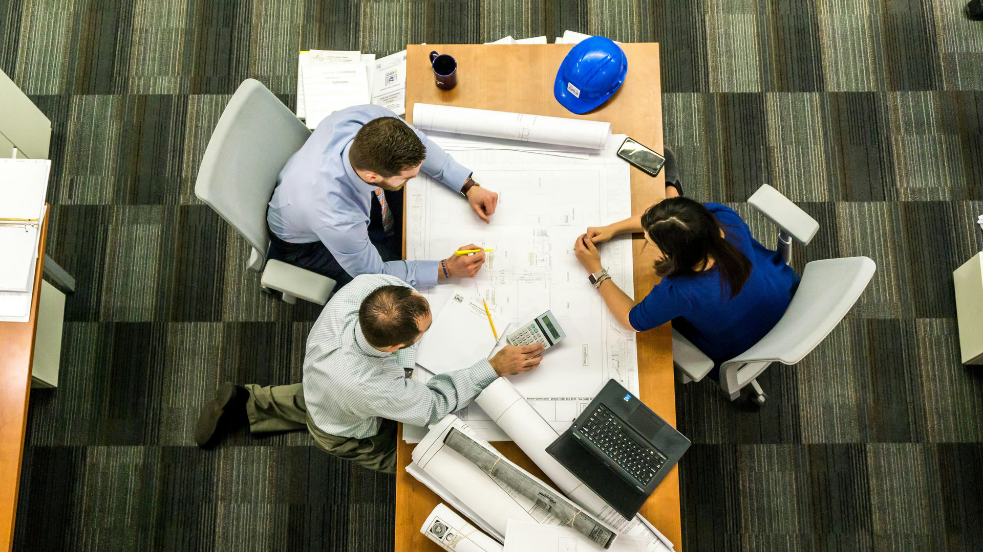 три людини сидять за столом і роздивляються папери з кресленнями про будівництво, на столі лежить синя каска, ці люди ріелтори