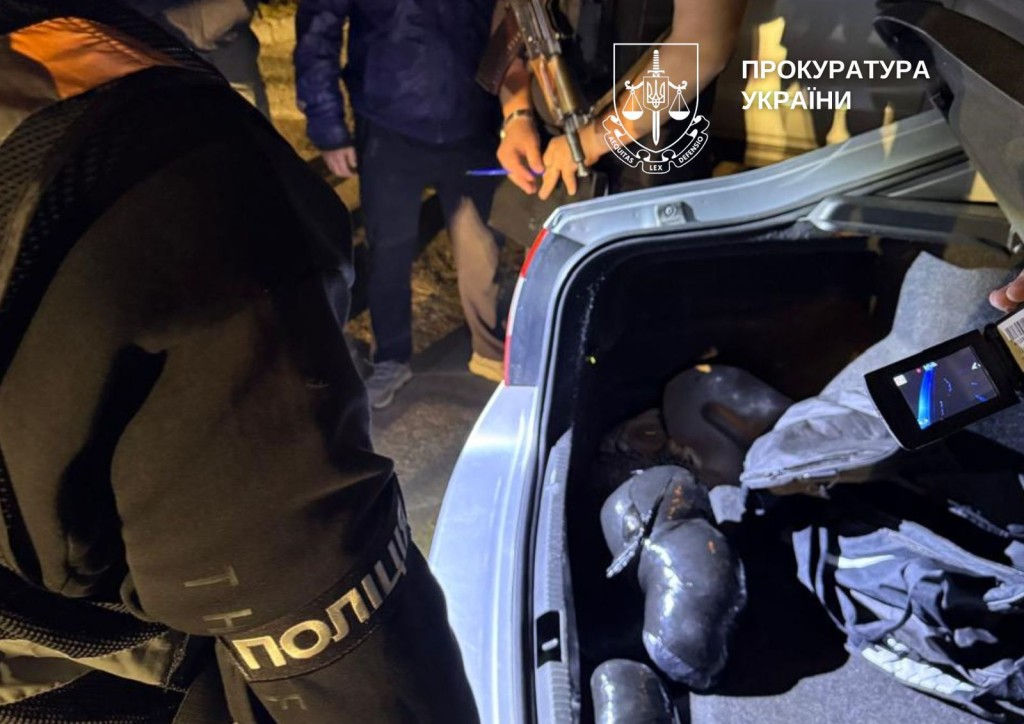 Чоловік із надписом Поліція на рукаві куртки стоїть біля відкритого багажника автомобіля, його колега фільмує це на камеру