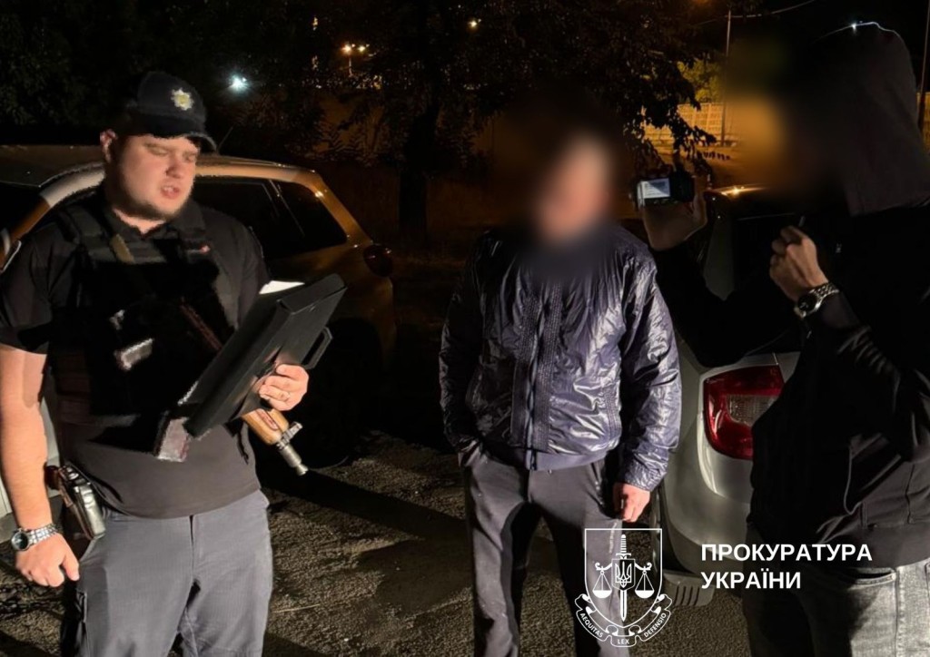 Вночі на вулиці працівник правоохоронних органів з автоматом на грулях зачитує з паперу підозру для затриманого чоловіка, що стоїть поруч