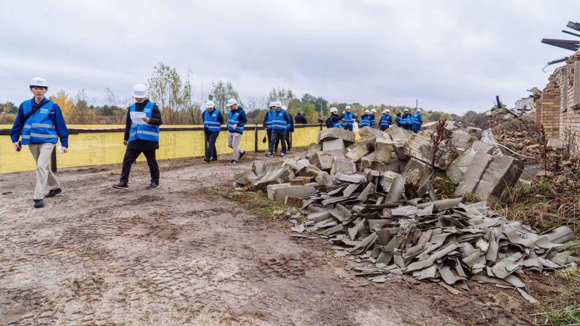 Чоловіки та жінки в синіх жилетах і касках ходять з-поміж зруйнованих будинків у приватному секторі