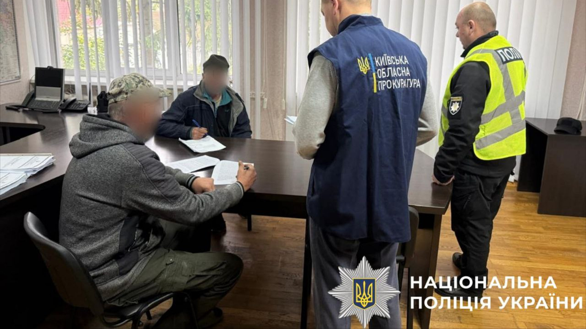 Двоє працівників поліції стоять біля столу, за яким сидять двоє підозрюваинх