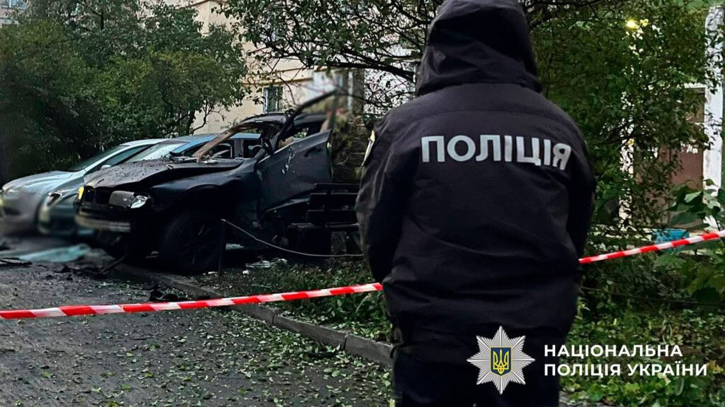 Працівник поліції стоїть біля червоної стрічки, яку повісили біля підʼїзду, де вибухнув автомобіль