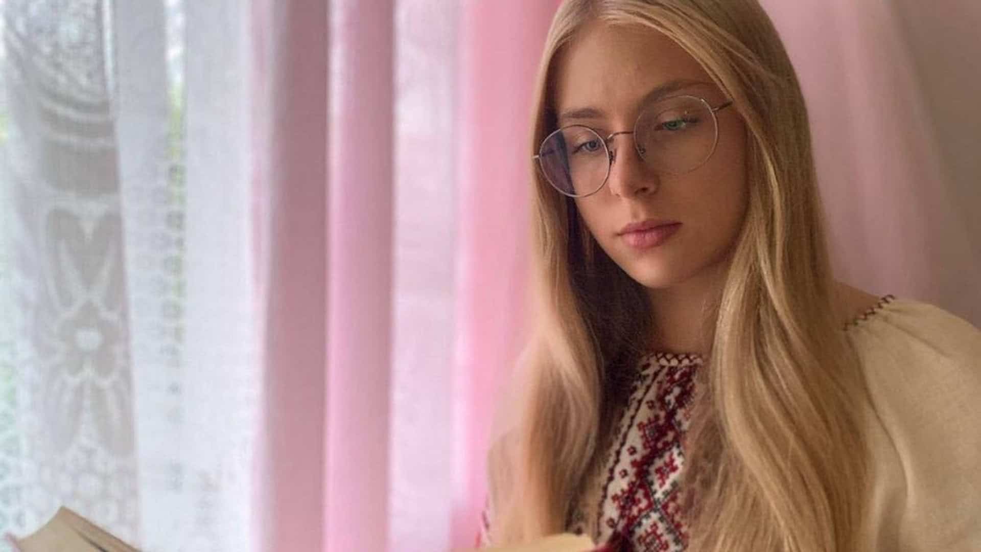 Біловолоса дівчина 18 років сидить на фоні рожевої кімнати і опустила очі в книжку, ця дівчина загинула під час атаки на Київ