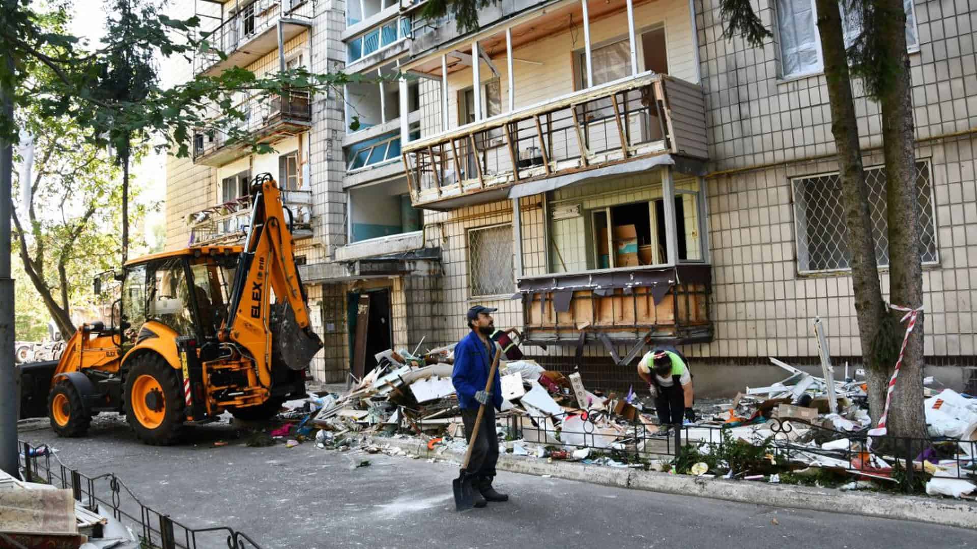 Подвірʼя багатоповерхового житлового будинку, уламки розбитих вікон лежать купою на асфальті, поряд працюють комунальники