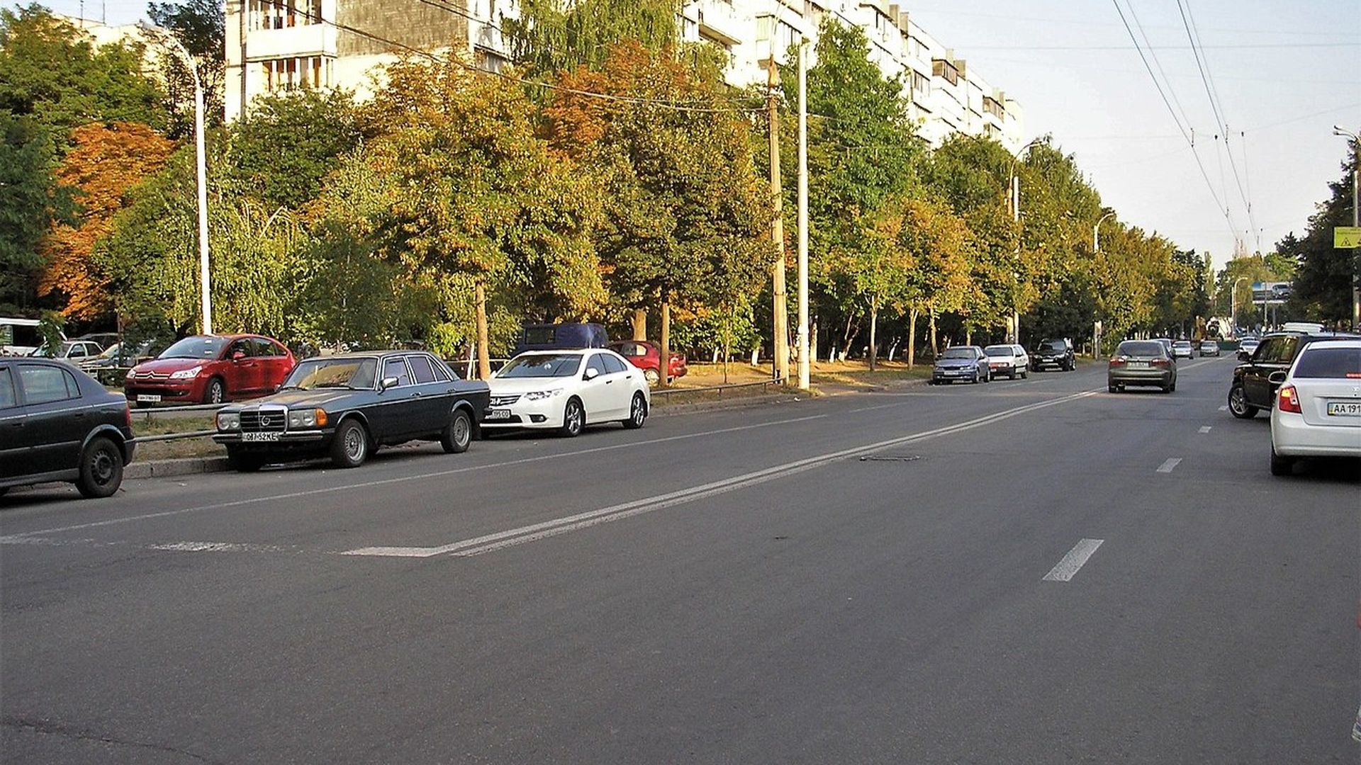 Міська вулиця з автомобілями обабіч дороги, житловими будинками та деревами вздовж тротуару