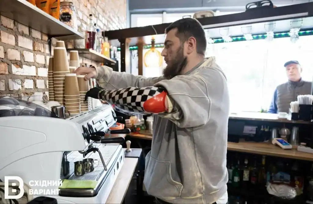 Ветеран з протезом руки варить каву на кавомашині, її замовив відвідувач, він стоїть трохи позаду