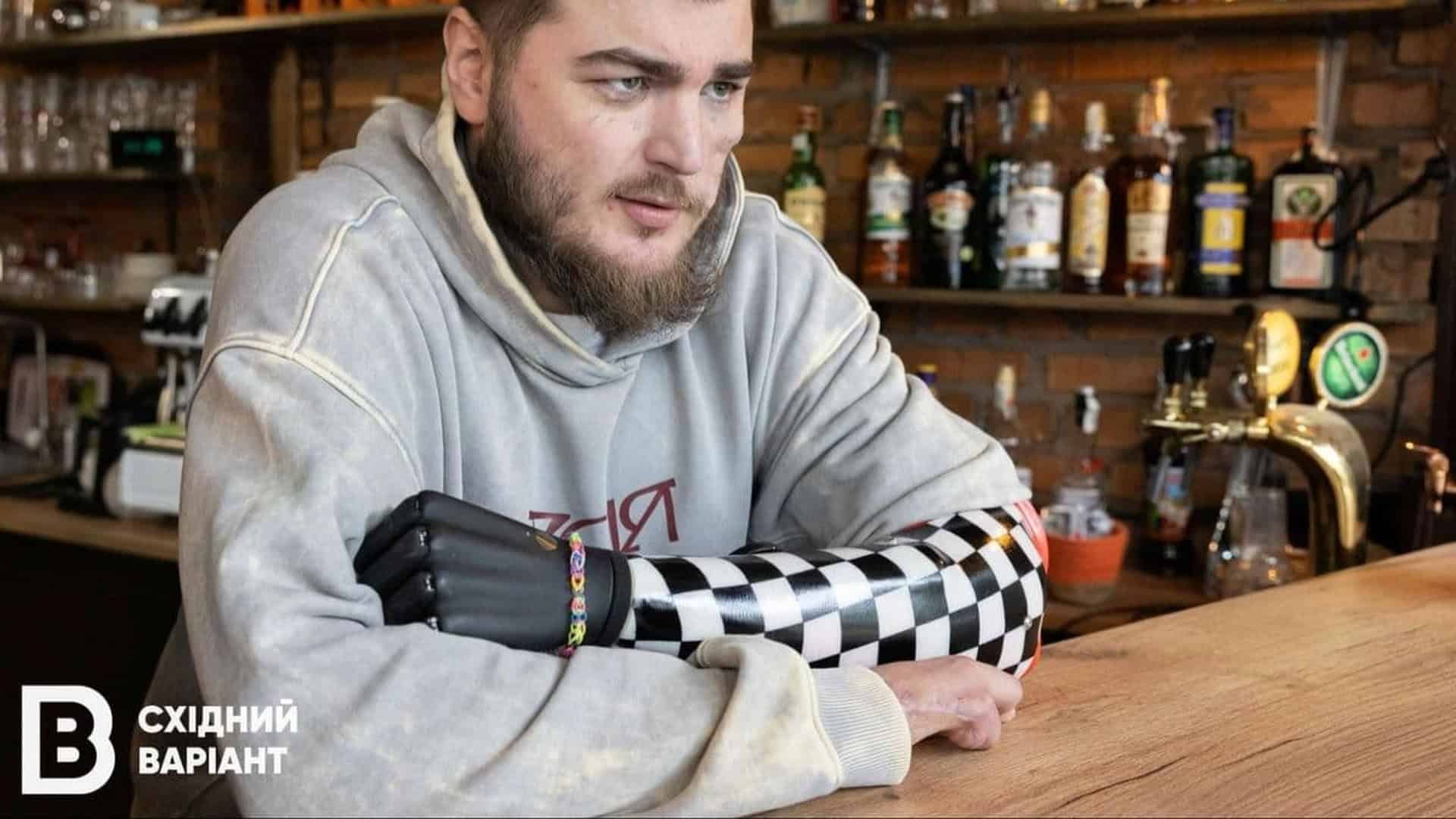 Ветеран із ампутацією руки стоїть за барною стійкою, позаду в нього бар кавʼярні
