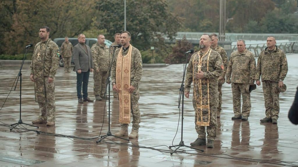 Київ: на Алеї Захисників відкрили пам’ятний знак воїнам ТРО України Військові капелани ТРО стоять на плацу на мокрій від дощу плитці перед мікрофонами