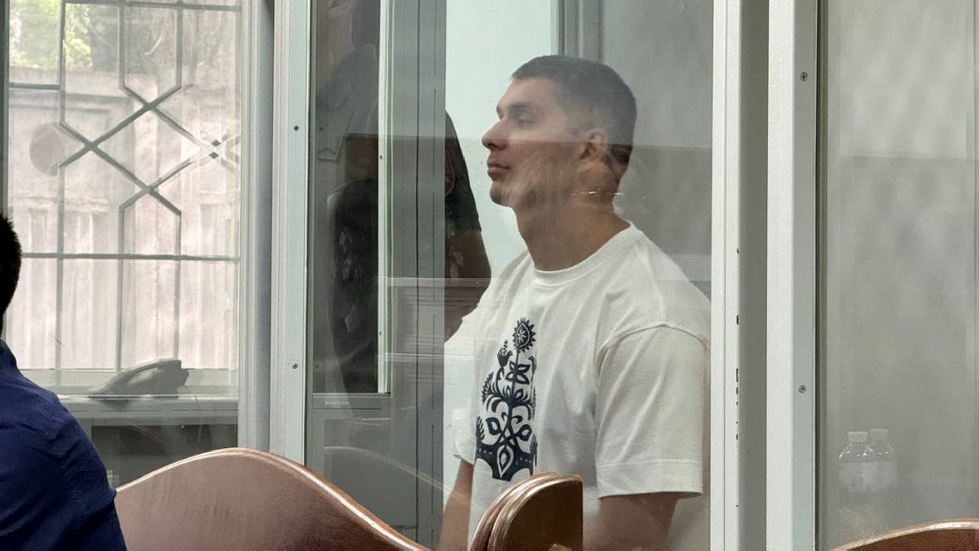 Чоловік у білій футболці сидить у скляній клітці під час судового засідання