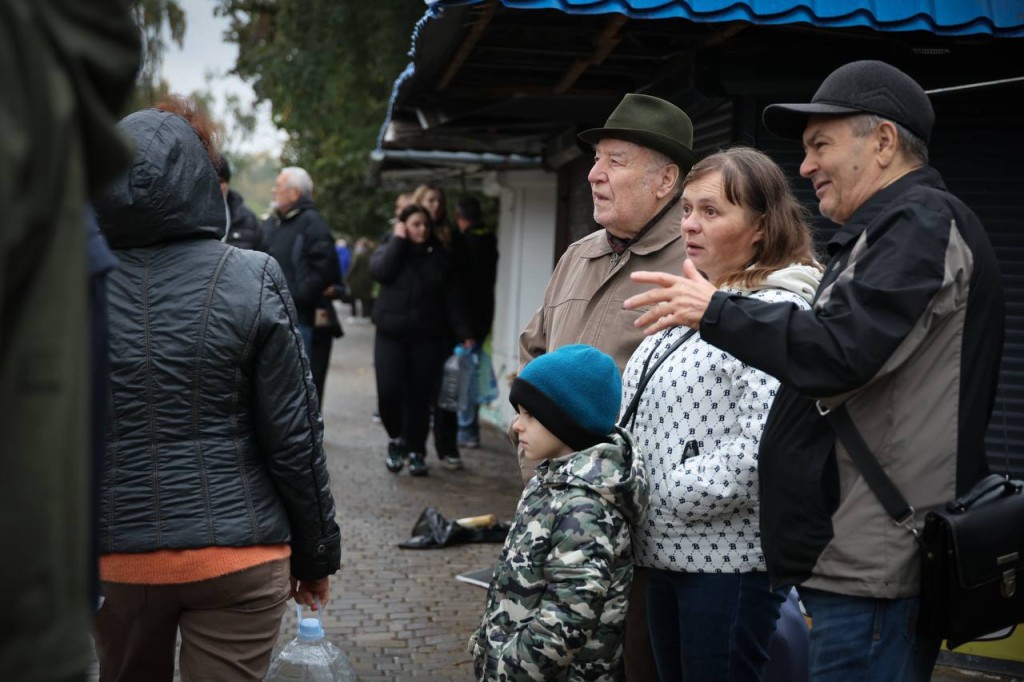 Місцеві жителі, серед них літні люди та дитина, стоять біля ринку, обговорюючи події