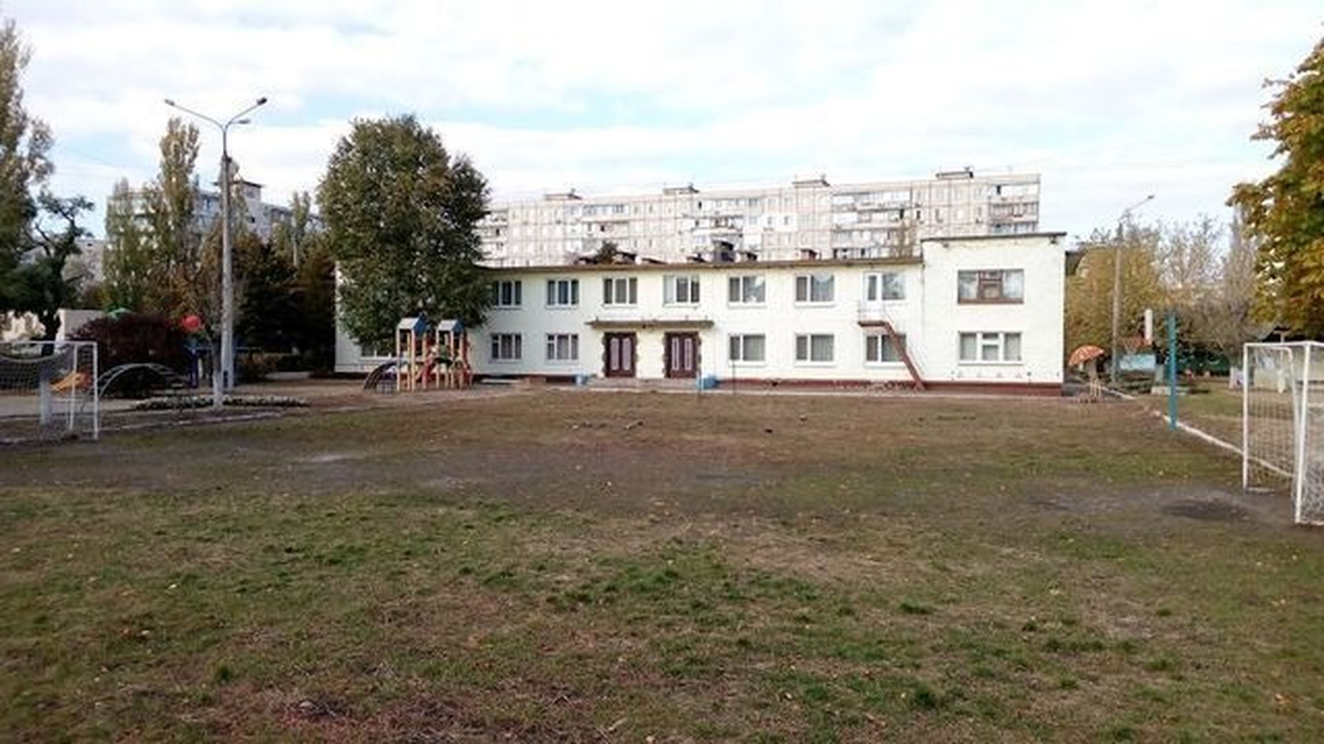Двоповерхова біла будівля дитячого садка з ігровим майданчиком та футбольним полем