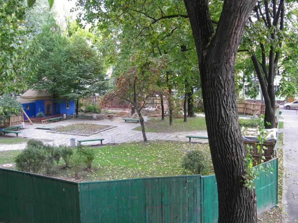 Попи Московського патріархату захопили сквер поруч зі штабом Сухопутних військ (фото) Зелений двір із деревʼяним парканом, лавками, синьою будівлею та великими деревами