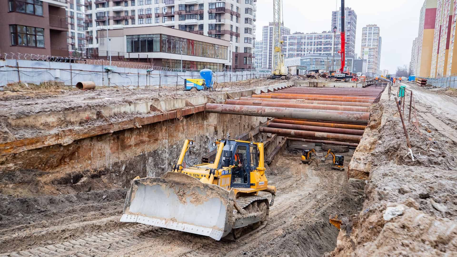 Будівництво метро на Виноградар, велика будівельна техніка працює в ямі на фоні нового житлового комплексу Варшавський квартал