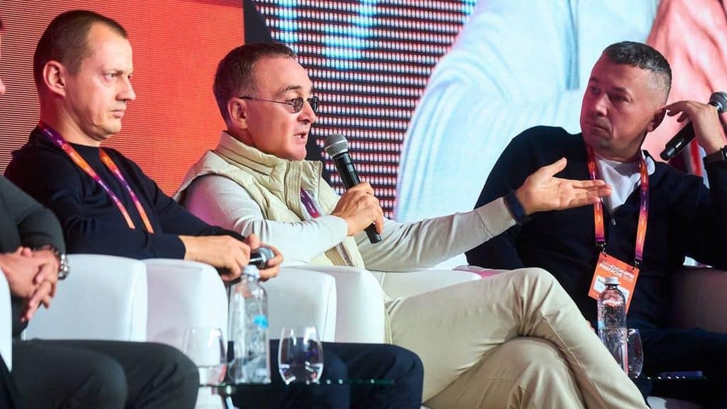 Нова квартира в Києві: прогноз подорожчання на 2026 рік Троє чоловіків сидять на стільцях на сцені, один з них з мікрофоном виступає на конференції житлових забудовників