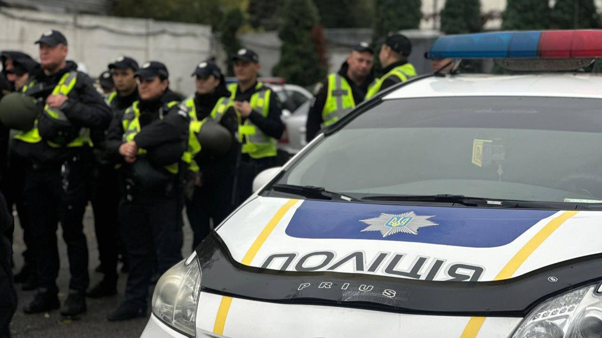 На передньому плані поліцейський автомобіль із увімкненим проблисковим маячком, на задньому — група поліцейських