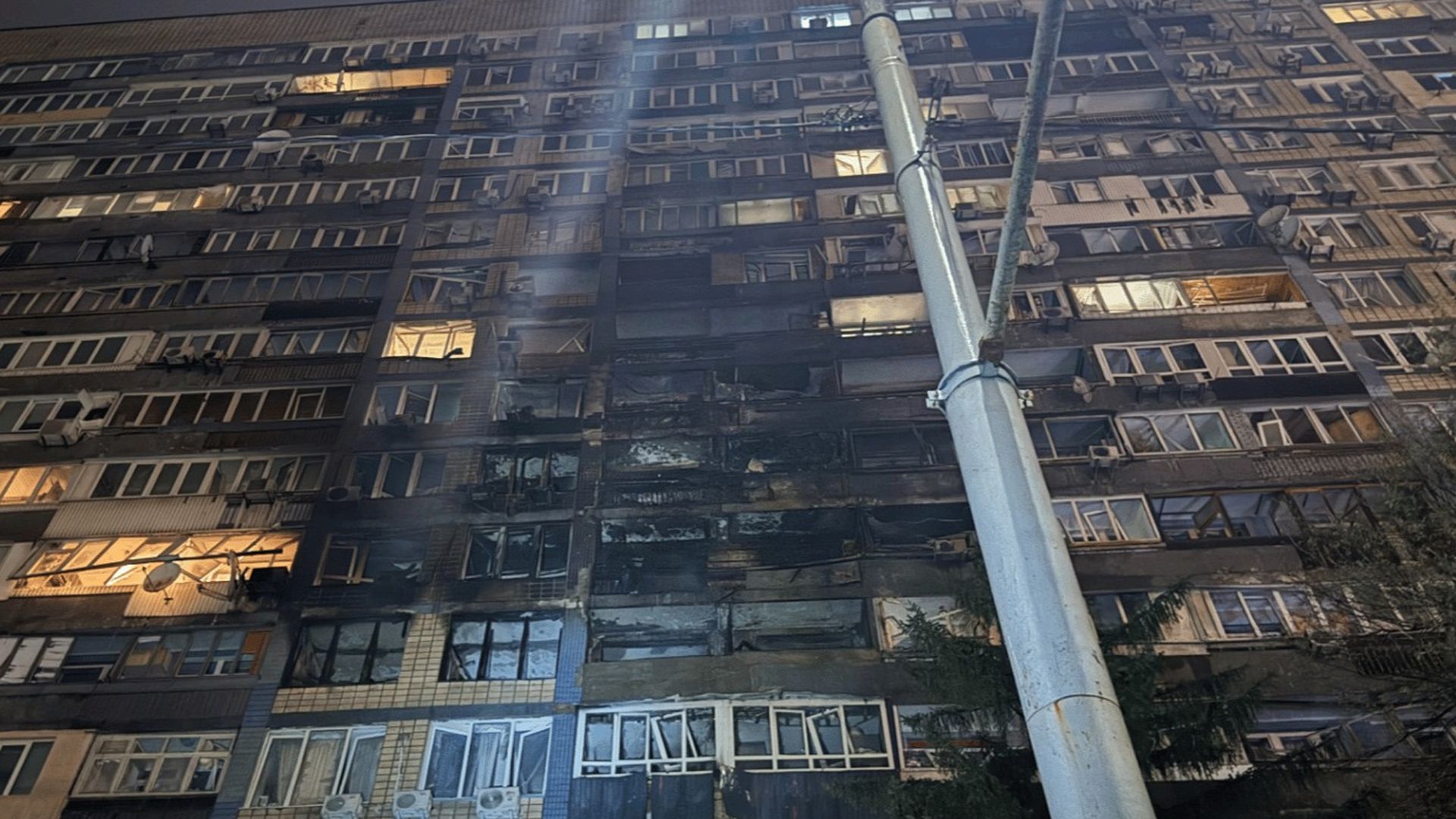 Висока багатоповерхова будівля з пошкодженими вікнами та слідами пожежі, знімок зроблено вночі