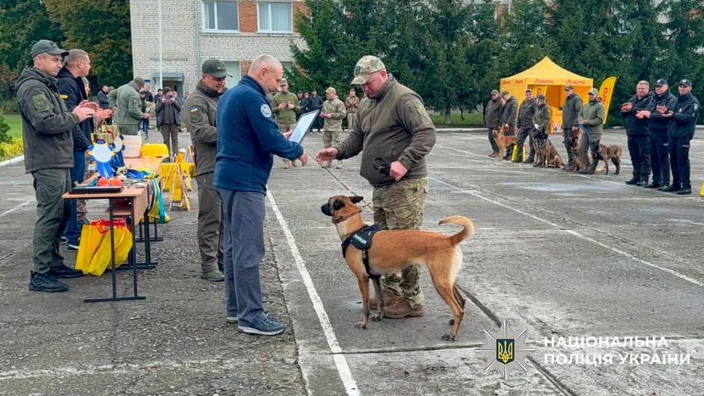 Псу поліцейському і його тренеру вручають нагороду на плацу, біля них ще кілька учасників