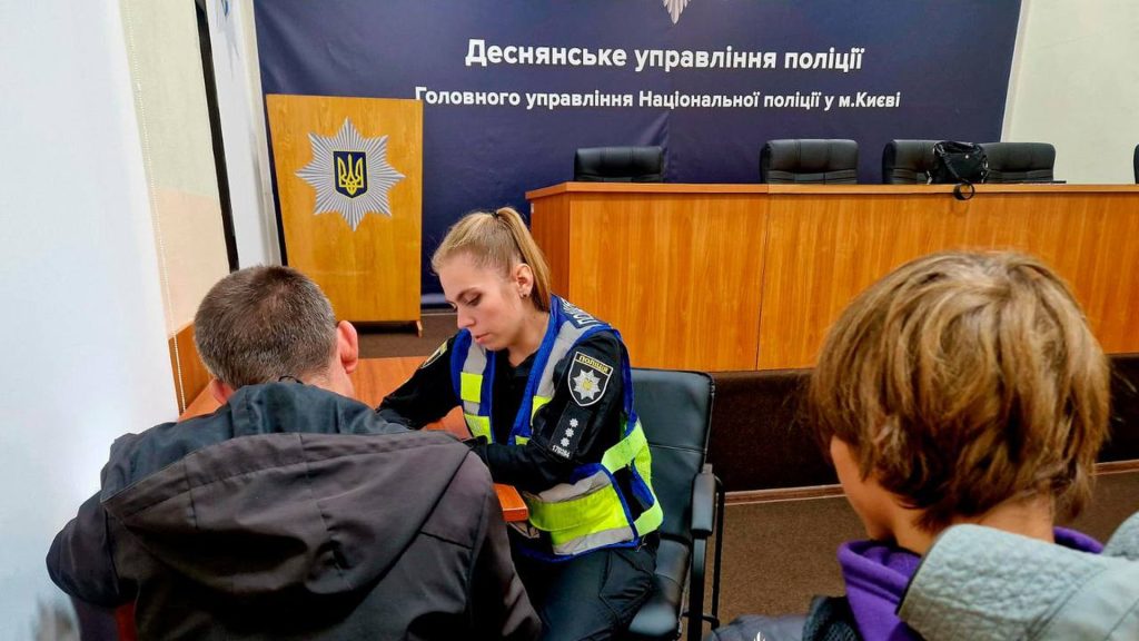 Офіцер поліції сидить за столом і дивиться на документи на столі, поряд із нею сидять двоє чоловіків