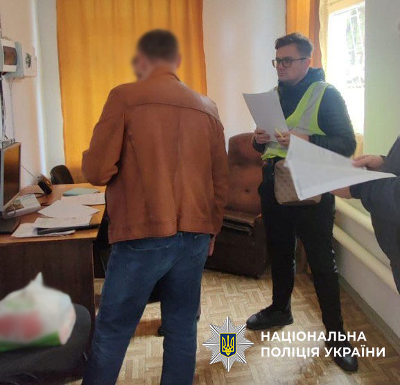 Працівник поліції в окулярах і з документами стоїть перед чоловіком в шкіряній оранжевій куртці, поряд ще кілька працівників поліції, вони говорять із затриманим