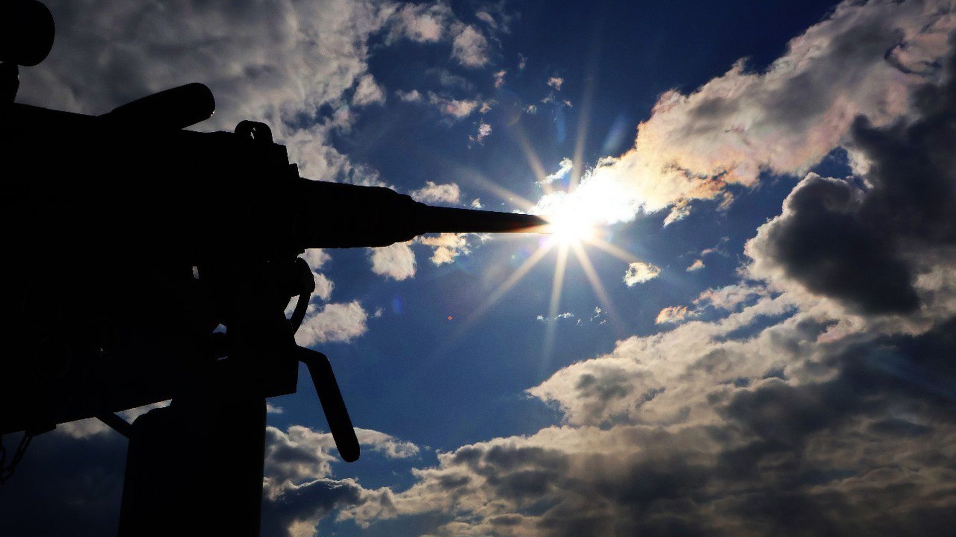 Силует кулемета спрямований у бік сонця, яке пробивається крізь хмари на яскравому денному небі