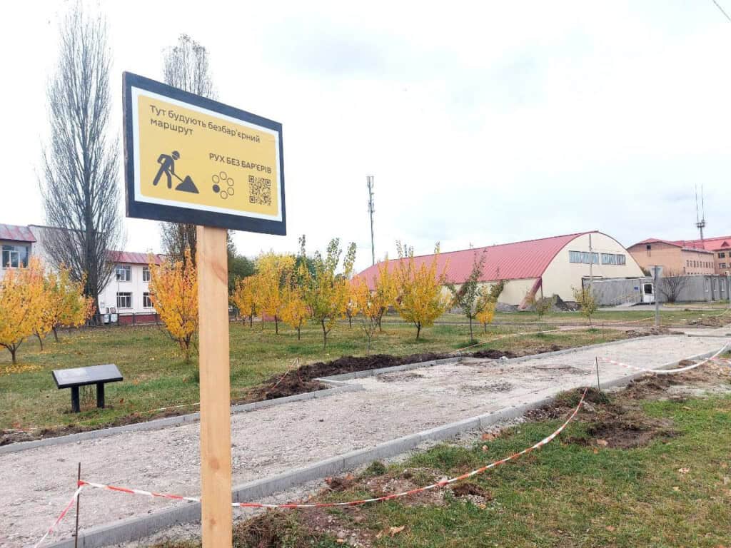 Тротуарна доріжка в стану будівництва в Славутичі на фоні жовтих дерев і таблички із надписом про безбарʼєрний простір