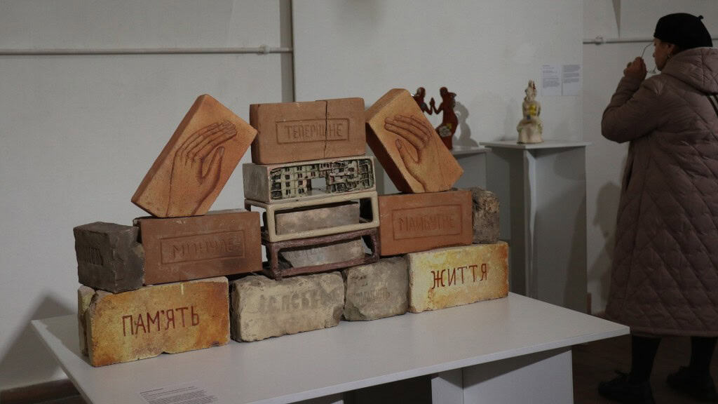 Цегла викладена на столі в мистецький витвір із різними надписами українською мовою: Мистецтво, памʼять, життя