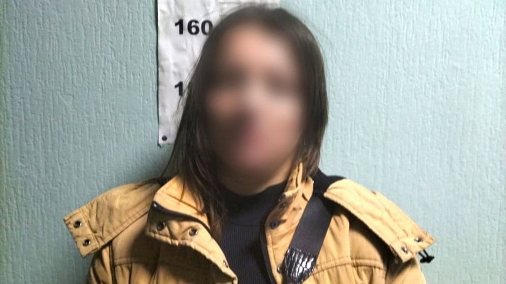 Жінка в жовтій куртці стоїть на фоні стіни в поліції із вимірювачем зросту, вона близько 160 сантиметрів