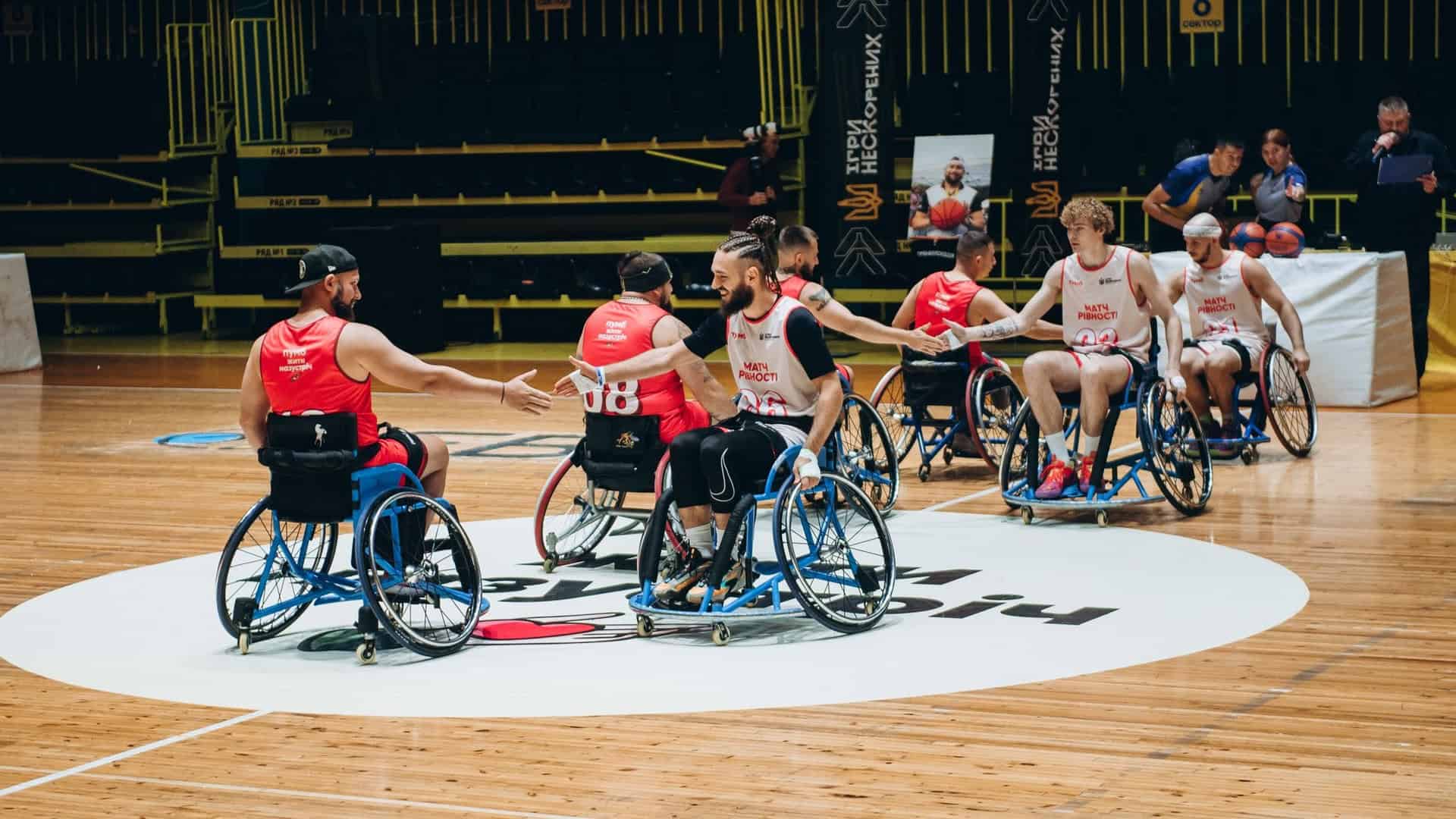Ветерани-спортсмени на кріслах колісних вітаються з командою одне одного на баскетбольному майданчику, на фото 6 людей, одна команда в білій формі, інша - в червоній