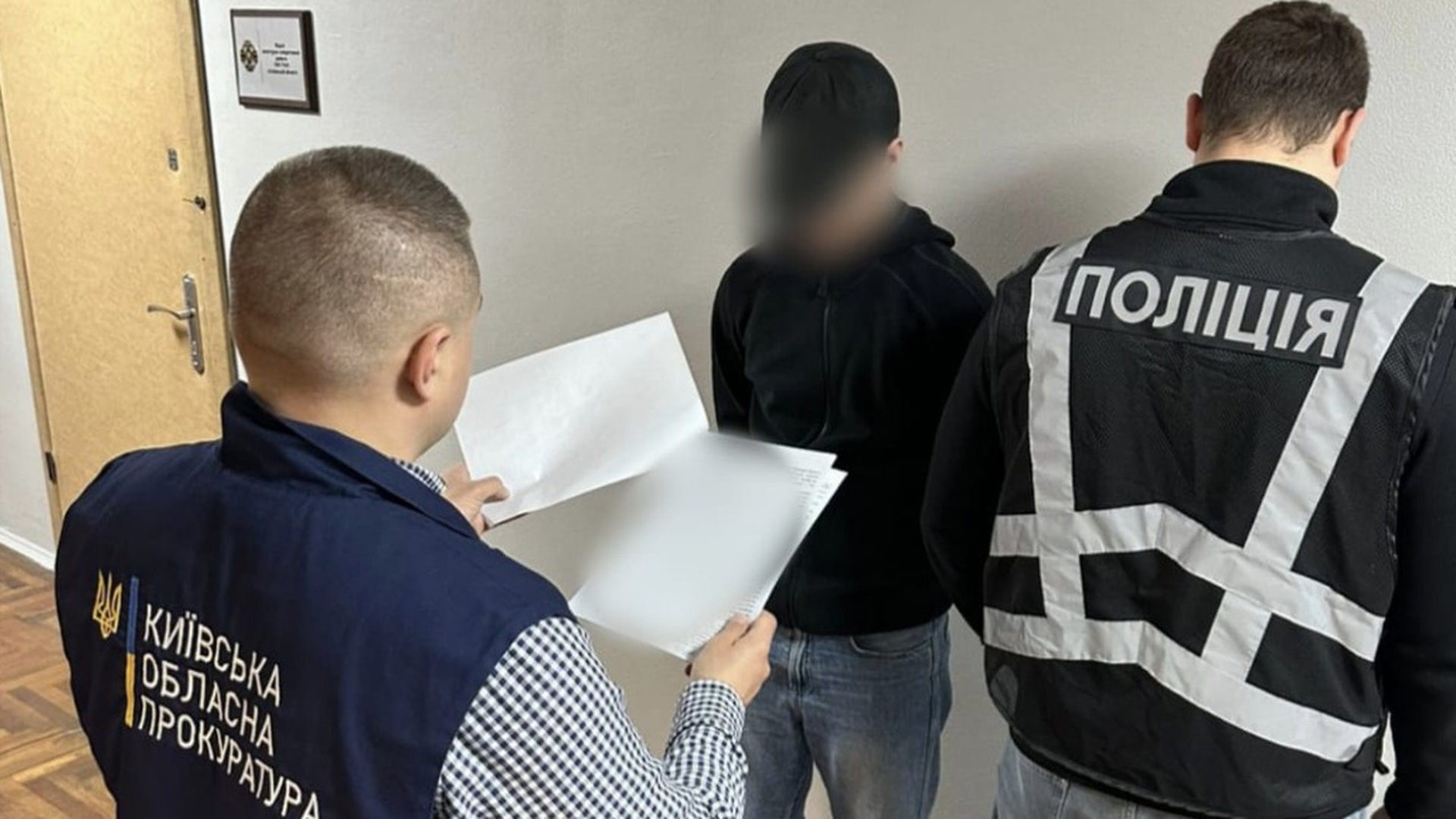 Працівник прокуратури зачитує щось з листків формату А4 підозрюваному, який стоїть в приміщенні біля стіни, біля підозрюваного також стоїть працівник поліції