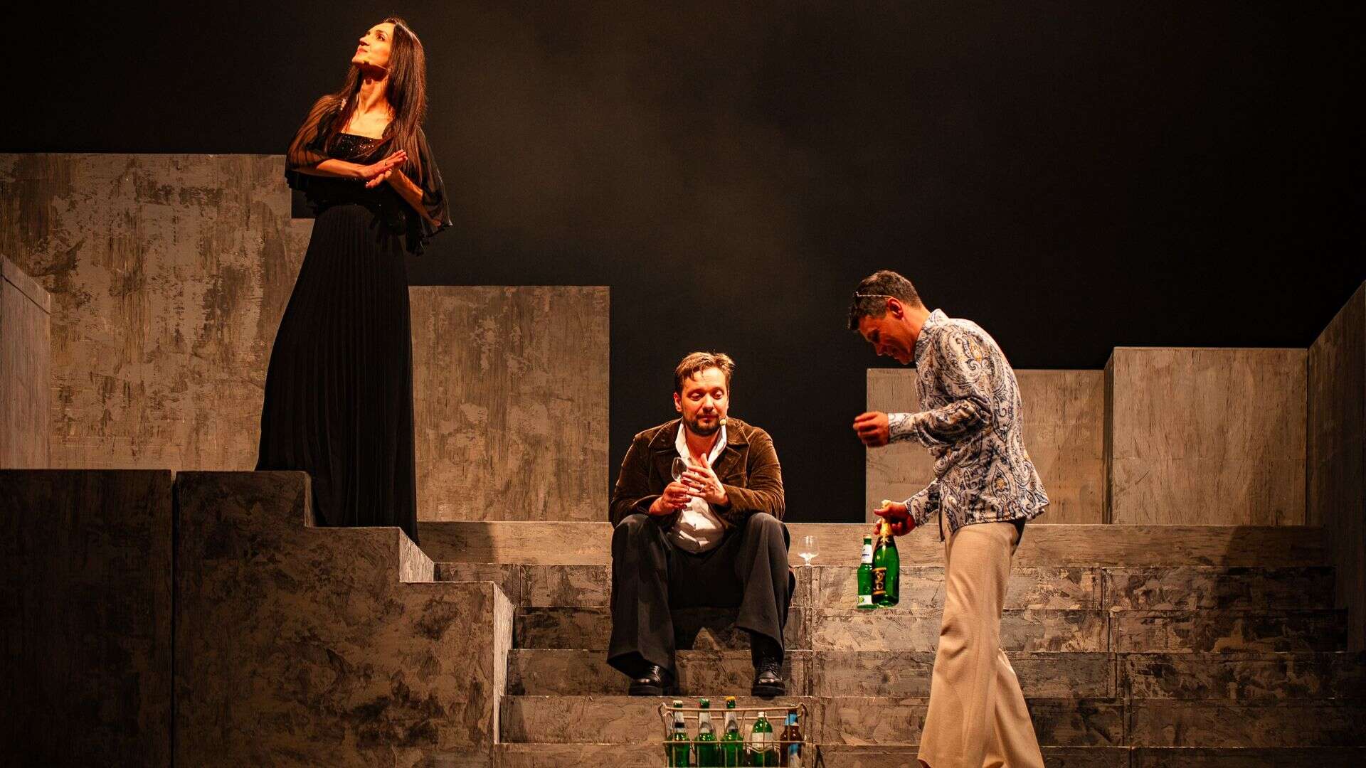 Троє акторів у виставі на сцені з декораціями у вигляді сірих сходів та кубів