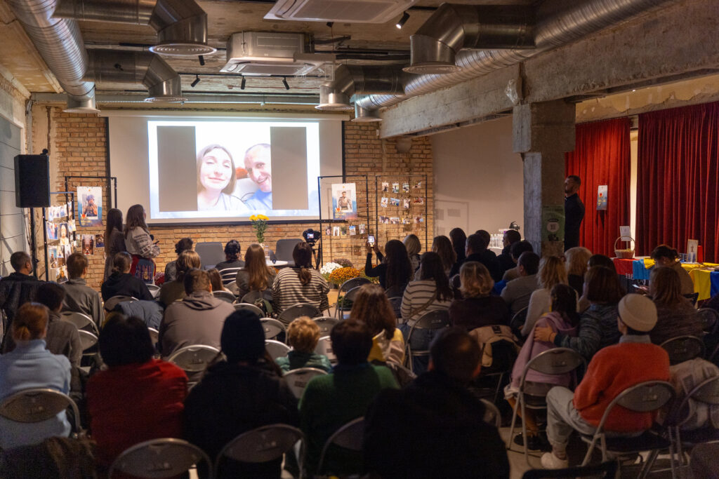 Фонд «Маємо жити» обʼєднав 26 міст України вечорами памʼяті загиблих захисників Захід у приміщенні: глядачі сидять у залі, на екрані демонструється фотографії пари