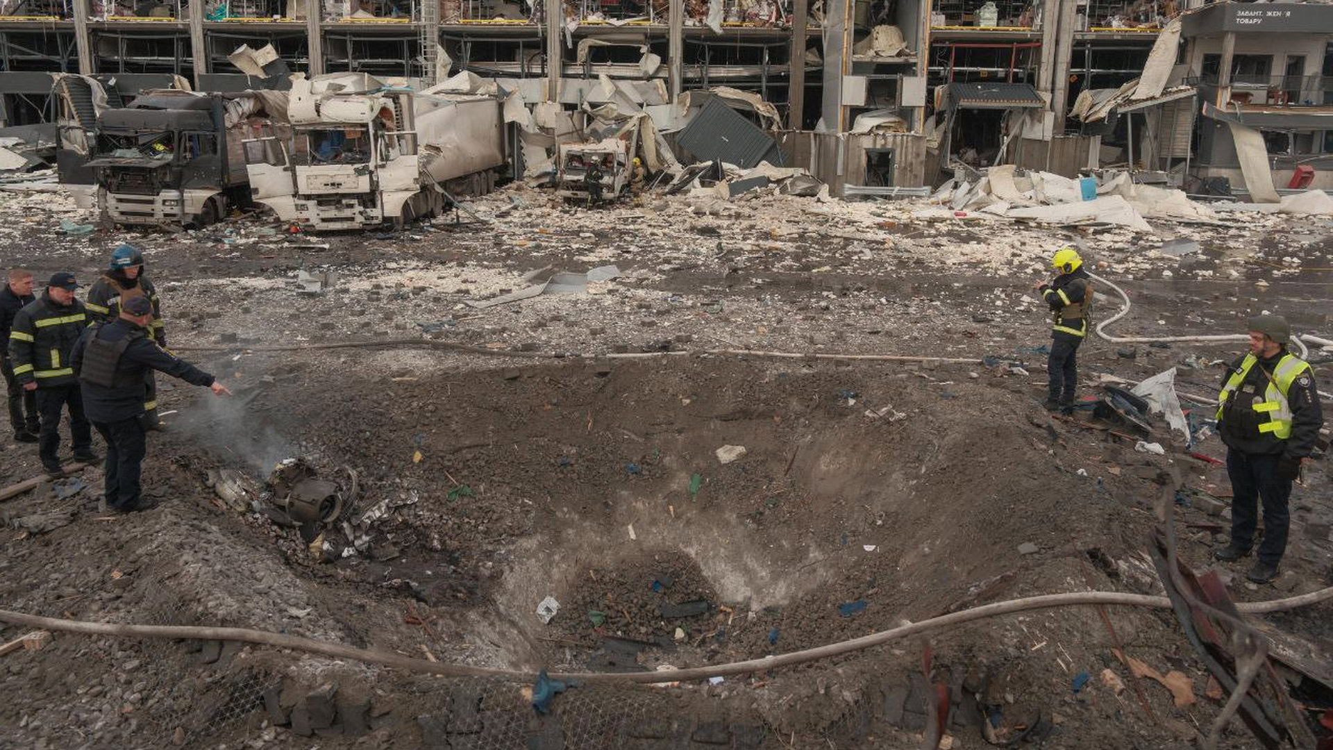 Величезна вирва в землі від удару ракети і поряд стоять люди, і видно знищене підприємство і фури