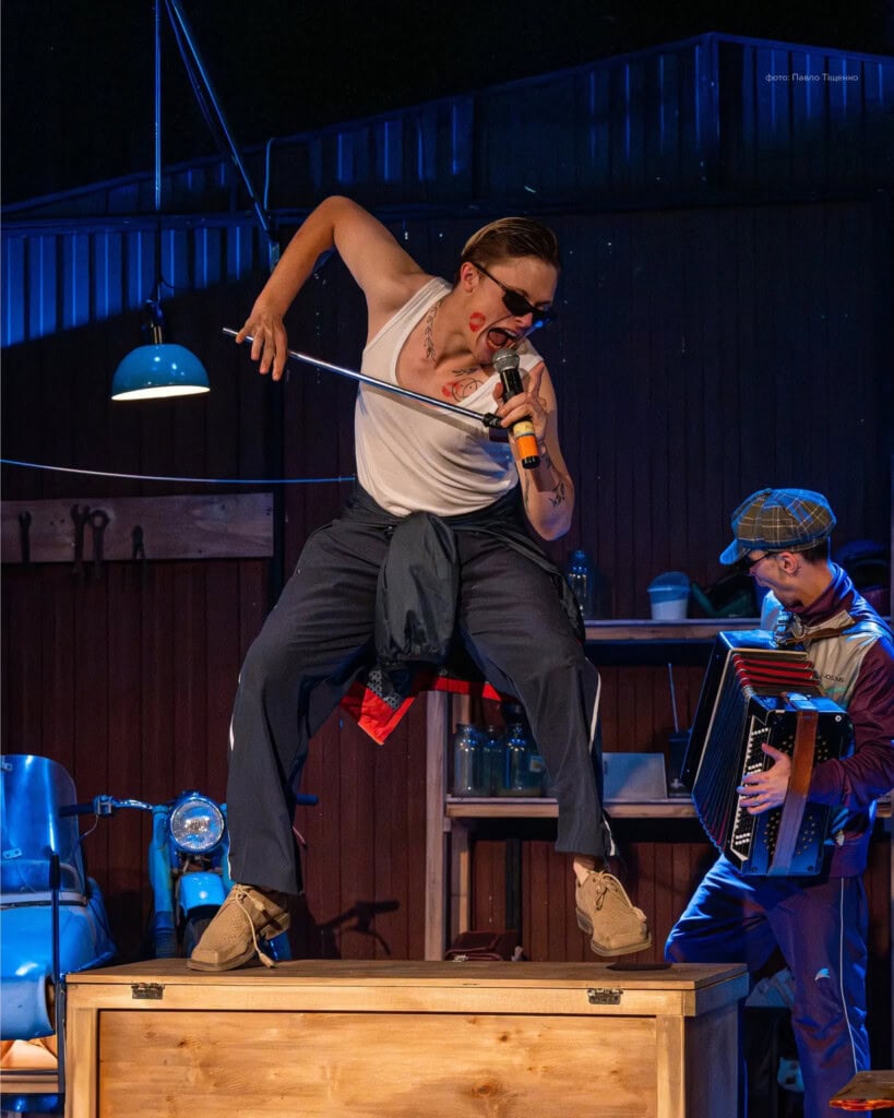 Актор у майці співає на ящику; музикант грає на баяні