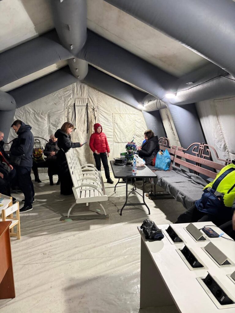 Люди в середині великого намету - тимчасового вокзалу, там є стільці і зарядні станції