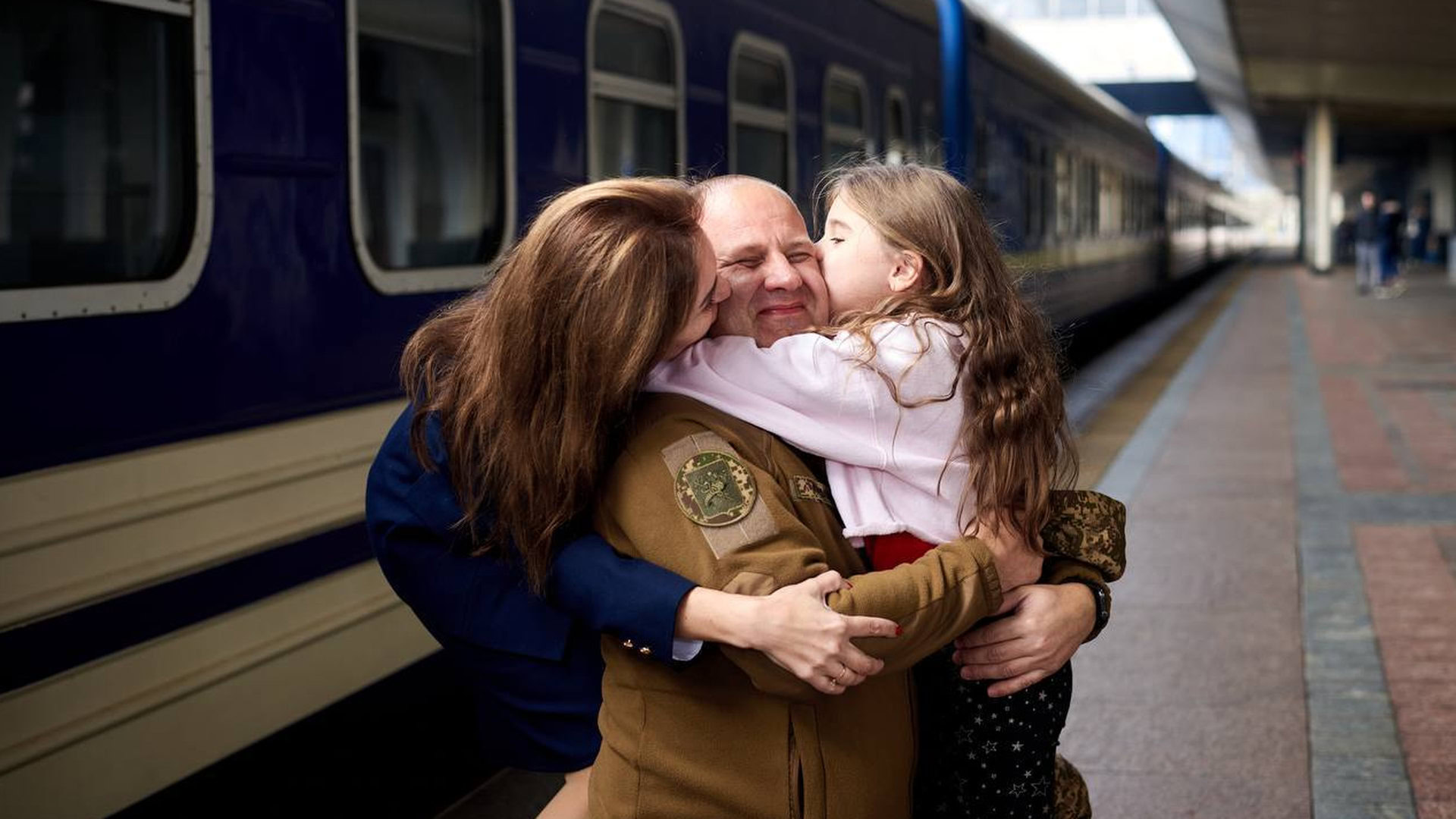 Донька і дружина обіймають військового на пероні вокзалу на фоні потягу