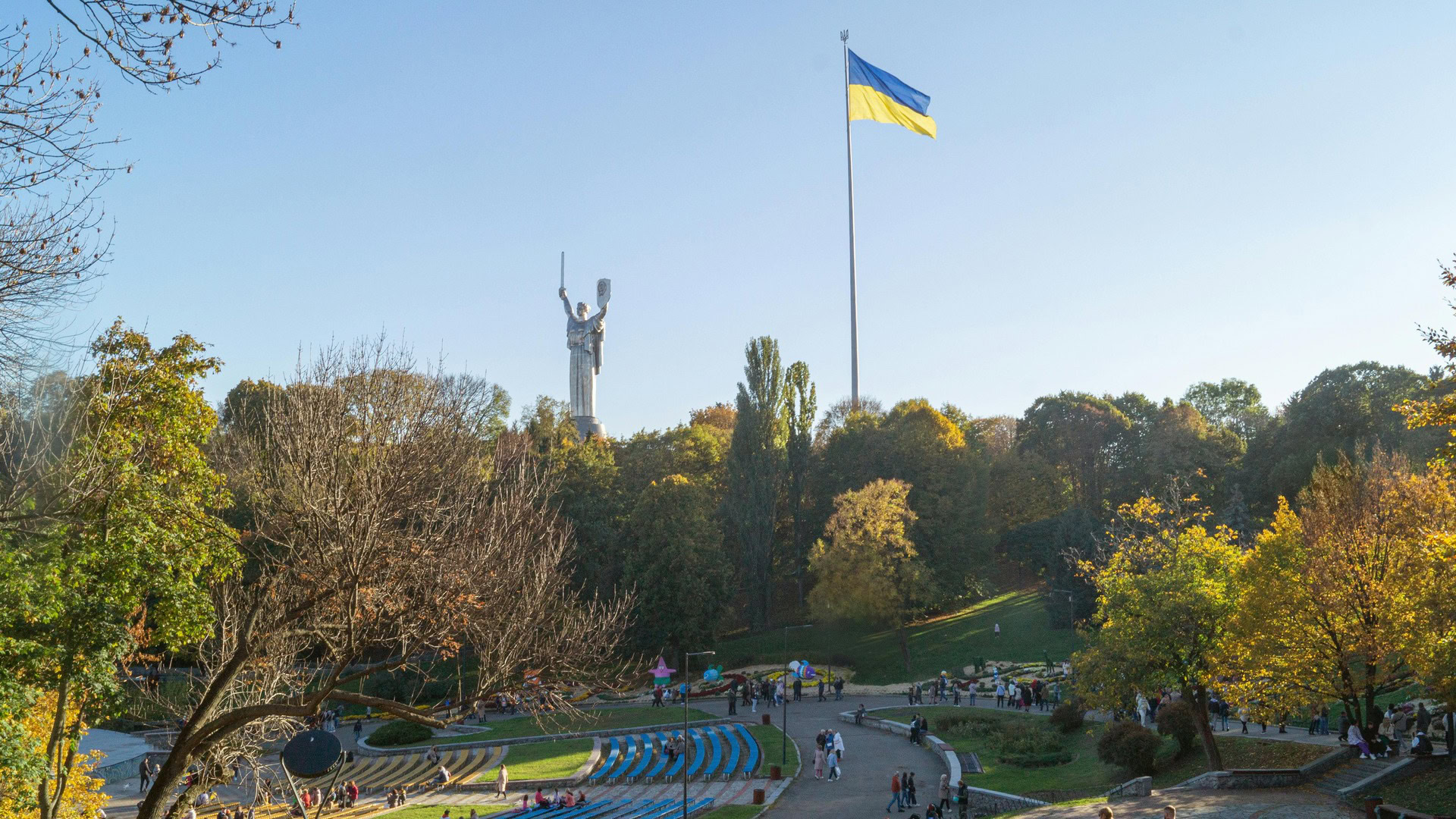 Осінній парк із прогулянковими алеями, монумент Батьківщина-Мати та великий прапор України на тлі блакитного неба в Києві