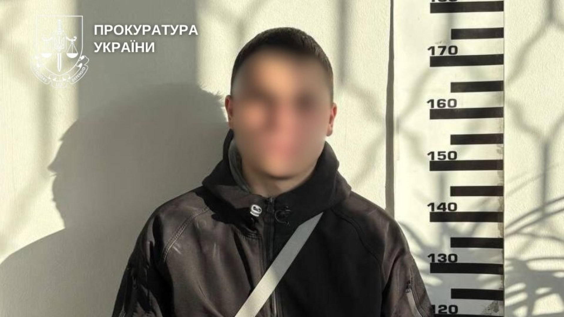 Підозрюваний у пособництві шахрайству з дронами стоїть біля ростоміра із заблюрованим обличчям у темній шкіряній куртці
