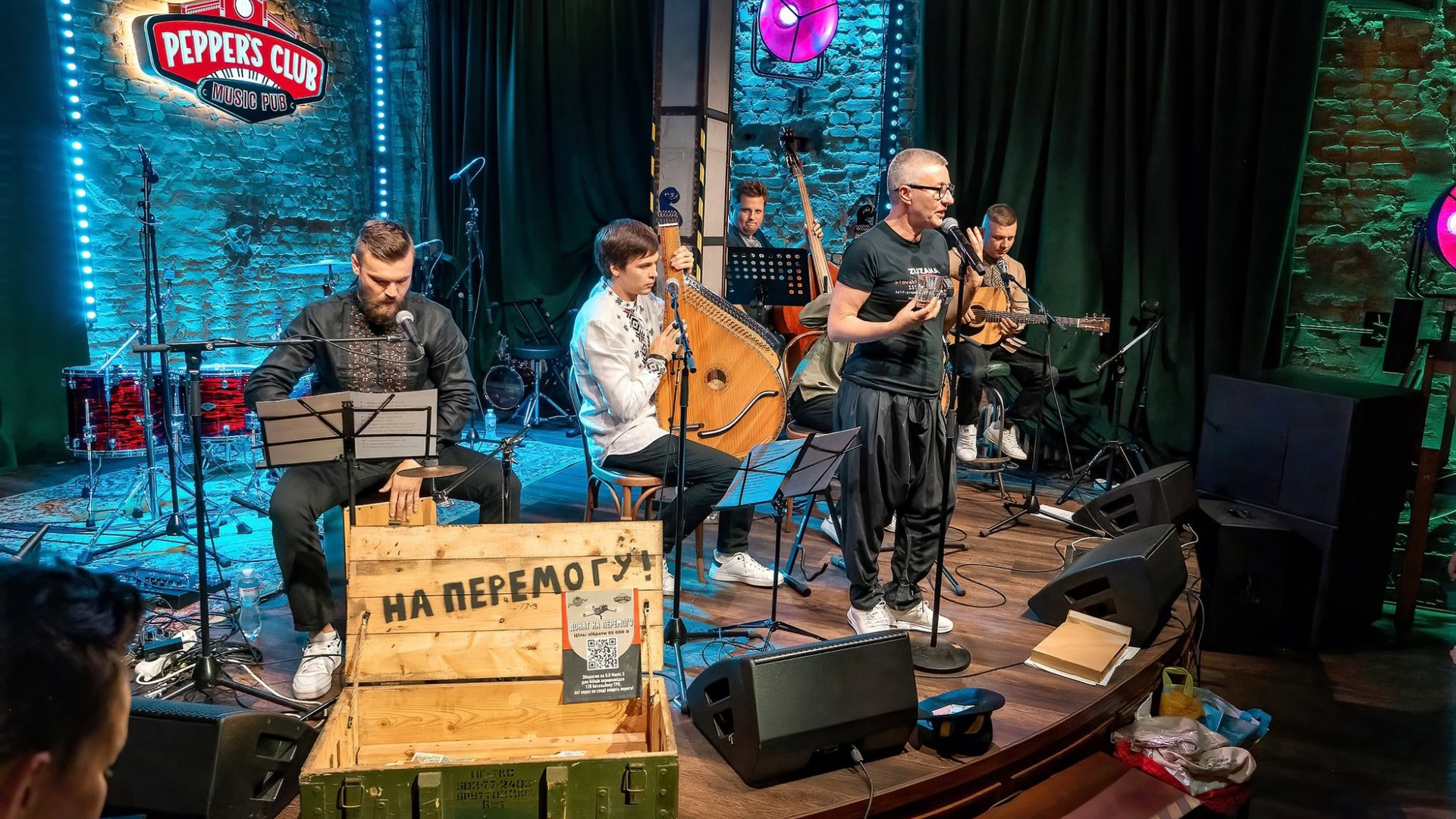 Музиканти виступають на сцені Peppers Club під час благодійного концерту «НаШапку», на передньому плані — ящик з написом «На перемогу!» для збору донатів