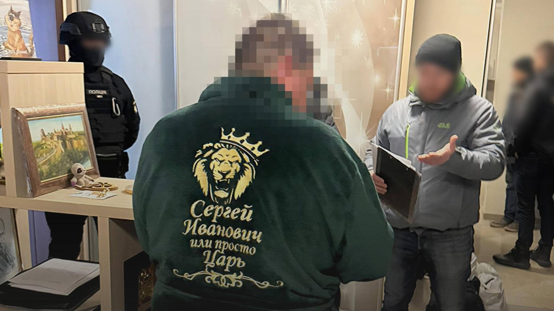 Підозрюваний у зеленому халаті з написом «Сергей Иванович или просто Царь» стоїть спиною до камери під час обшуку, поруч співробітники правоохоронних органів у формі