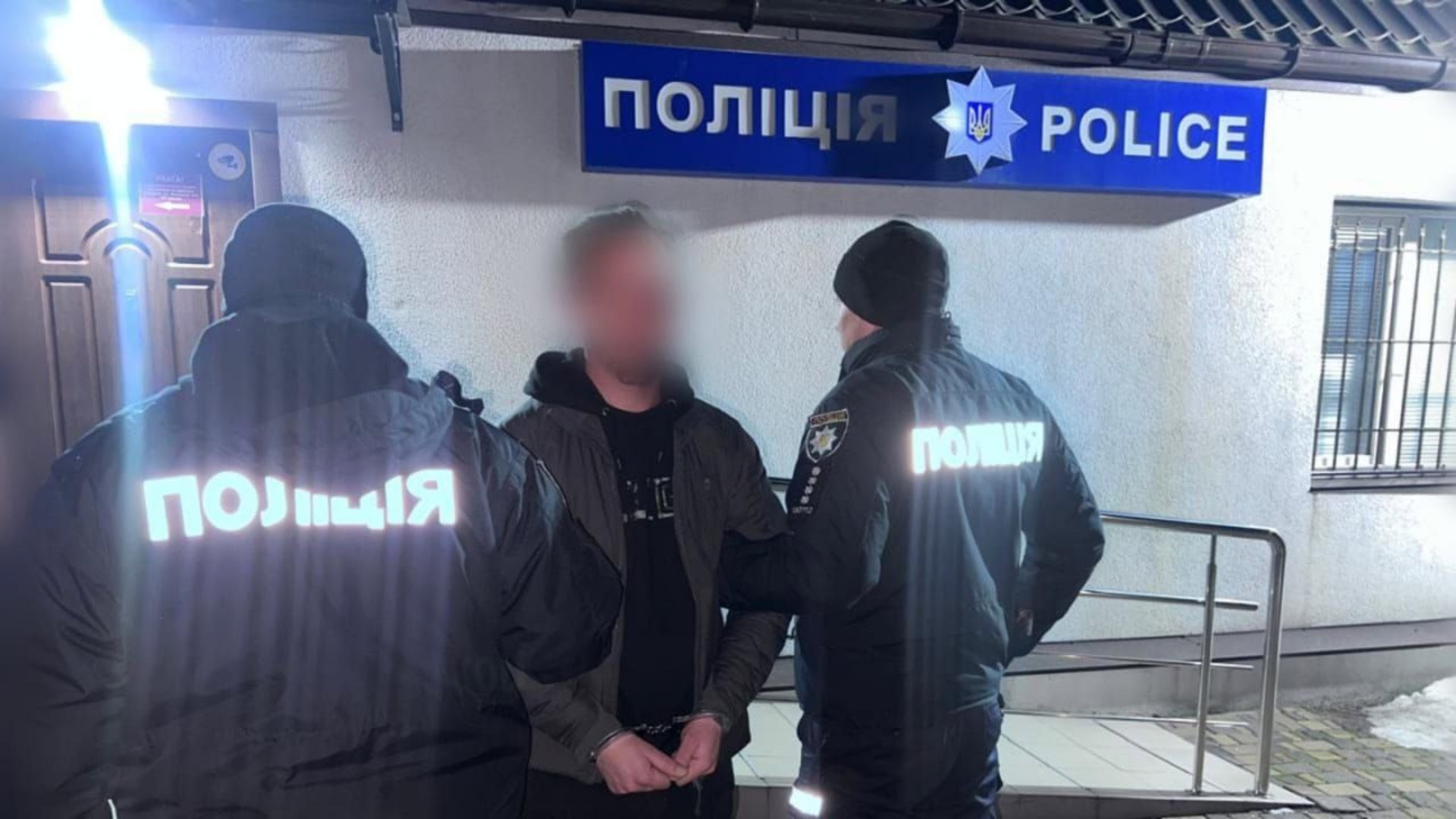 Двоє поліцейських з написом «Поліція» затримують підозрюваного з наручниками біля відділення поліції вночі