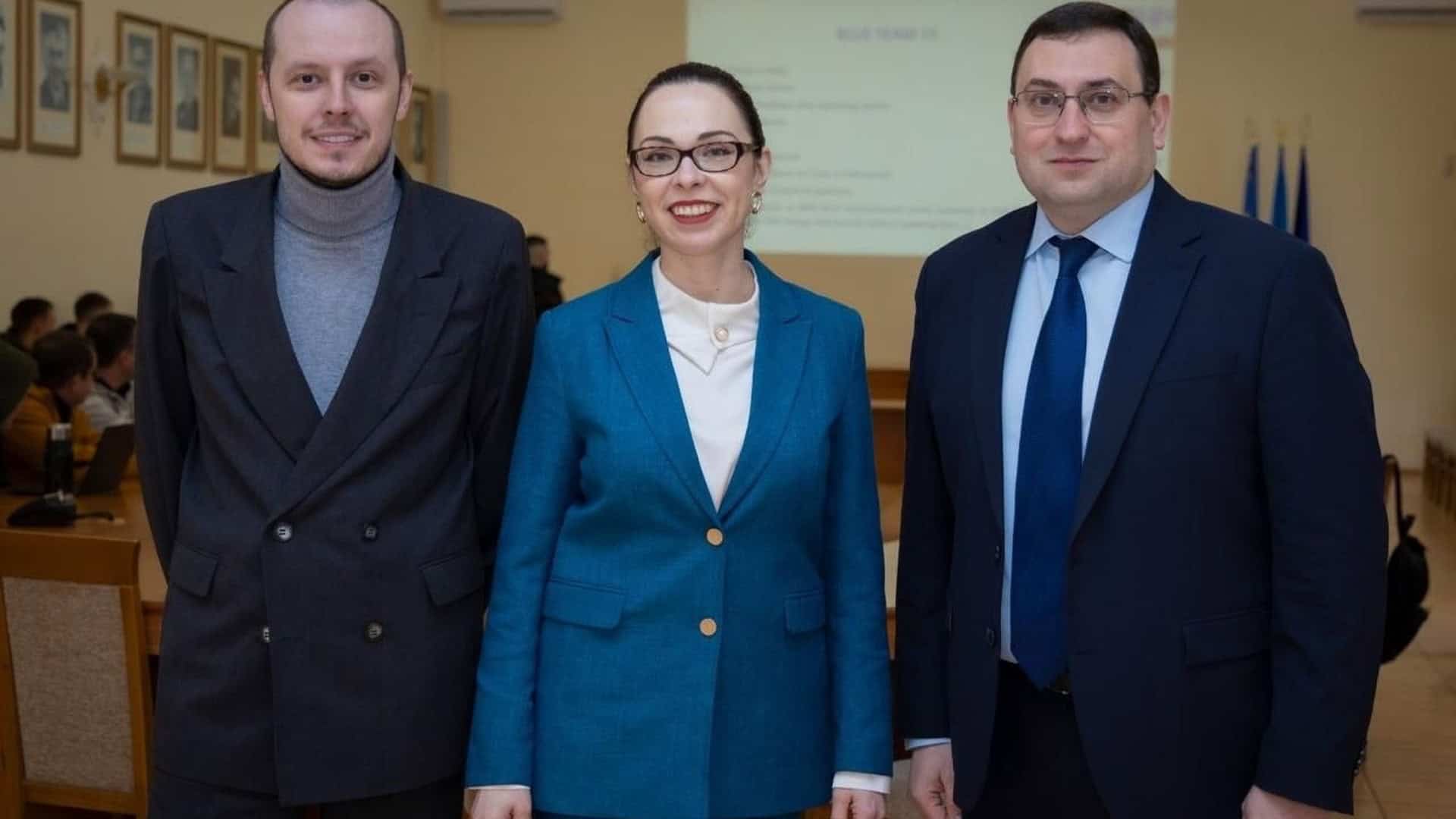 Троє учасників кібернавчань Locked Shields 2026 у КПІ ім. Ігоря Сікорського позують на фоні конференц-зали з презентацією та прапорами