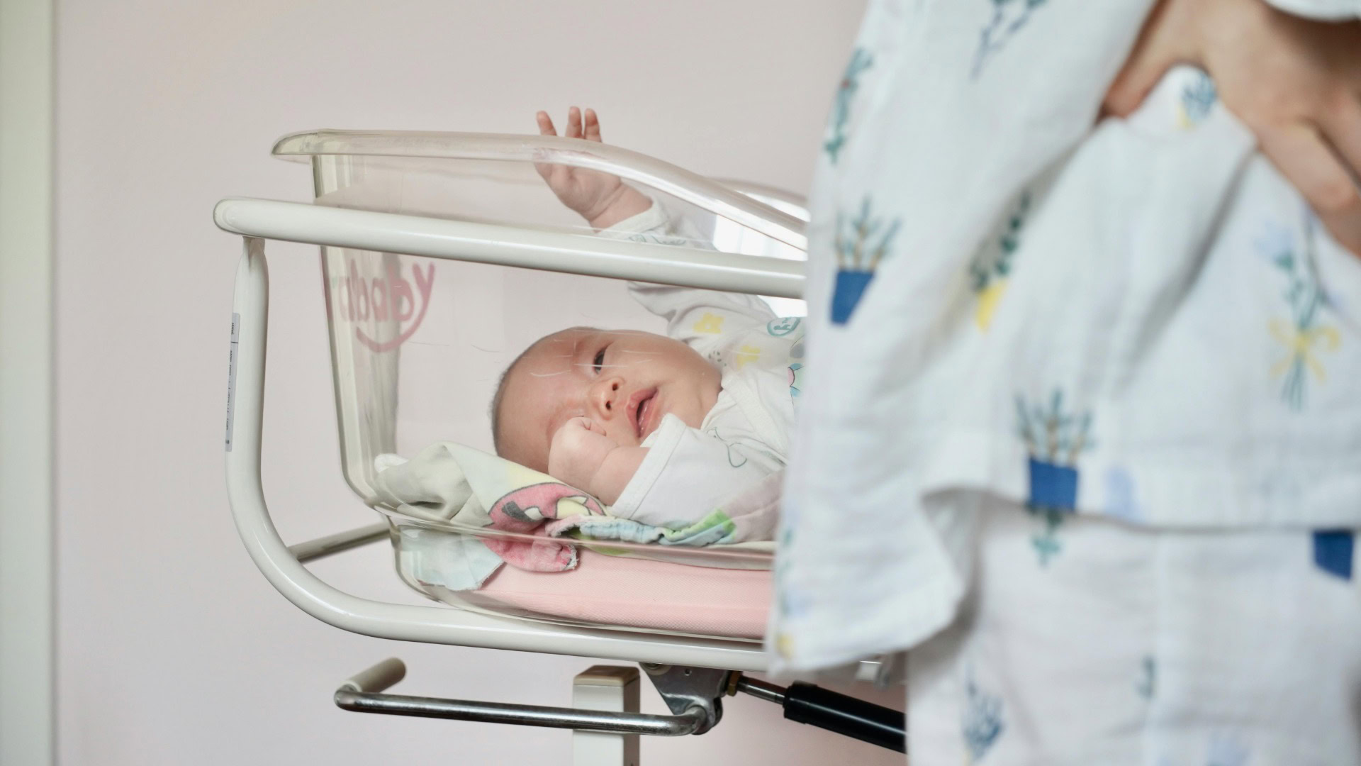 Новонароджений хлопчик лежить у ліжечку пологового відділення поруч із мамою у лікарняному халаті