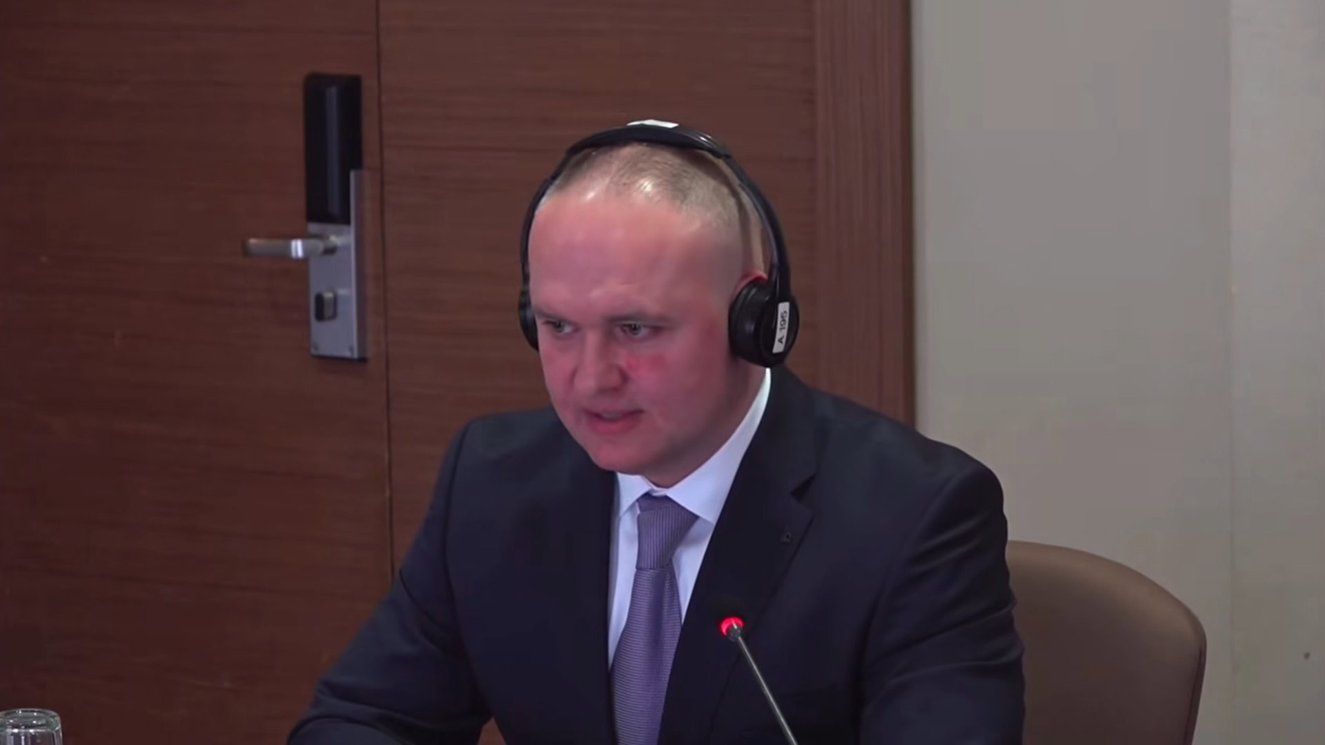 Орест Мандзій на засіданні в навушниках для перекладу виступає перед мікрофоном у діловому костюмі