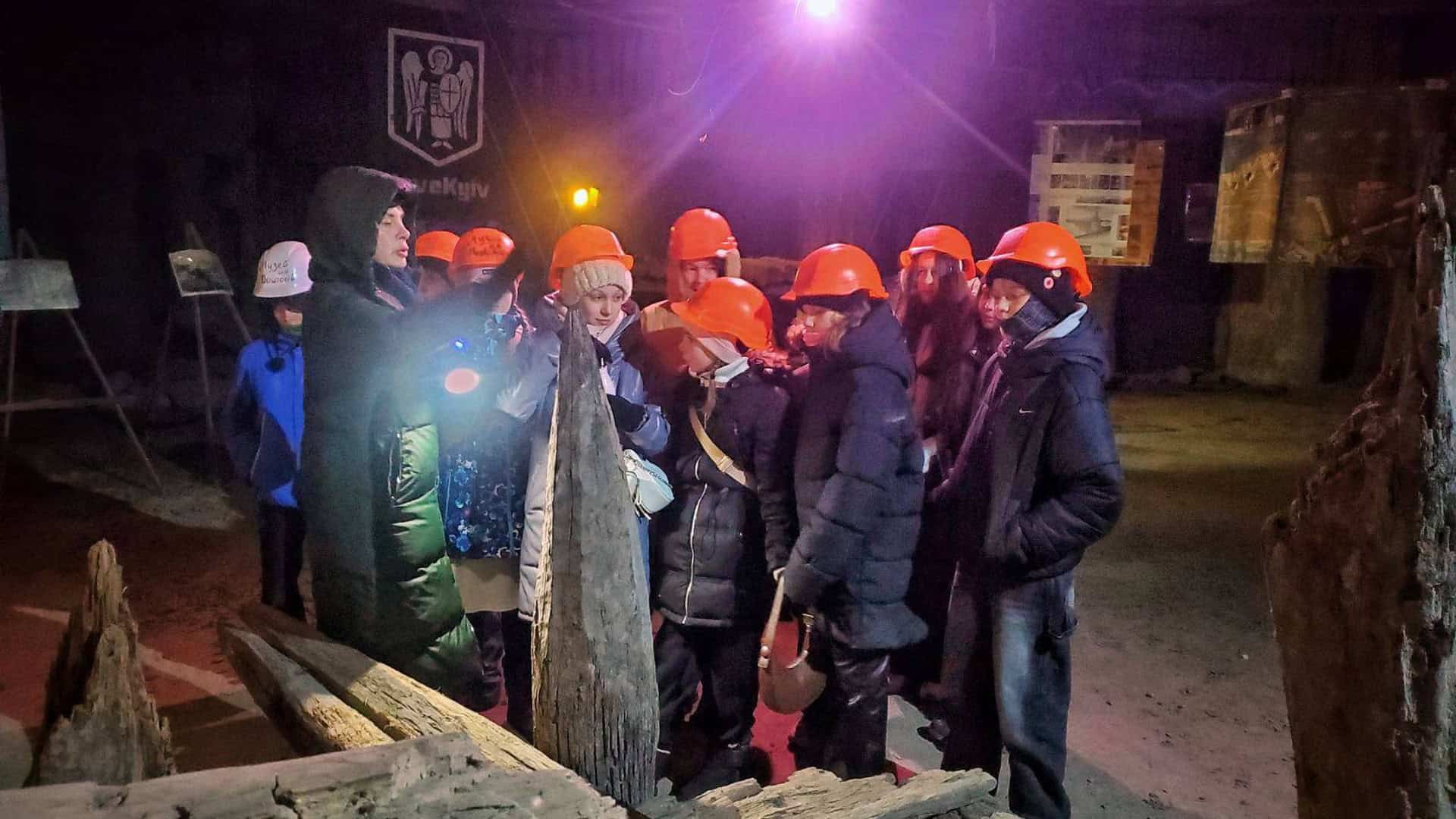 Діти в помаранчевих захисних касках та екскурсовод оглядають старовинні дерев'яні конструкції в підземній частині Музею на Поштовій площі у Києві