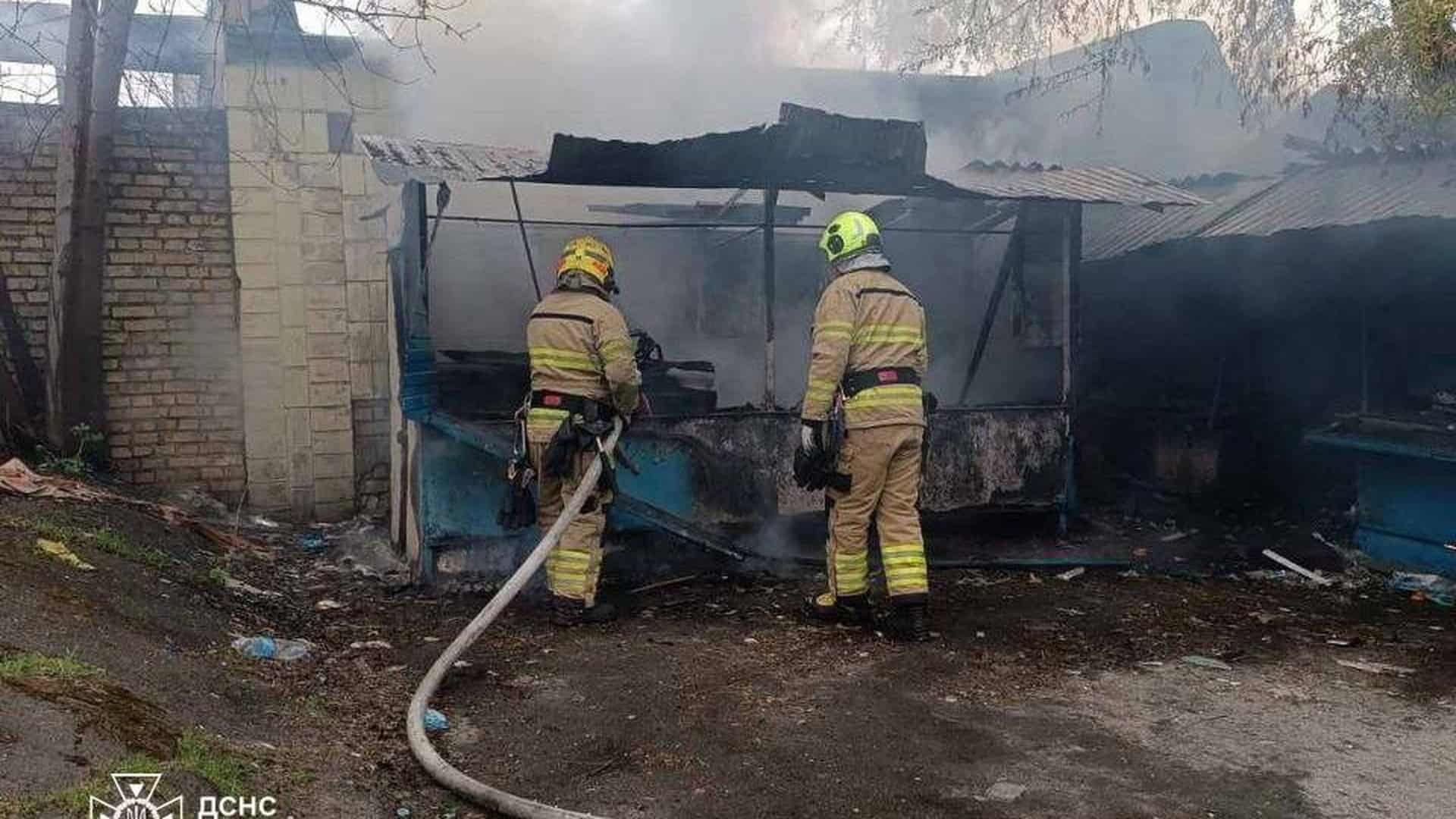 Рятувальники ДСНС гасять пожежу торгових кіосків на вулиці Симиренка у Святошинському районі Києва — двоє пожежників з рукавом на тлі диму та напівзруйнованих споруд