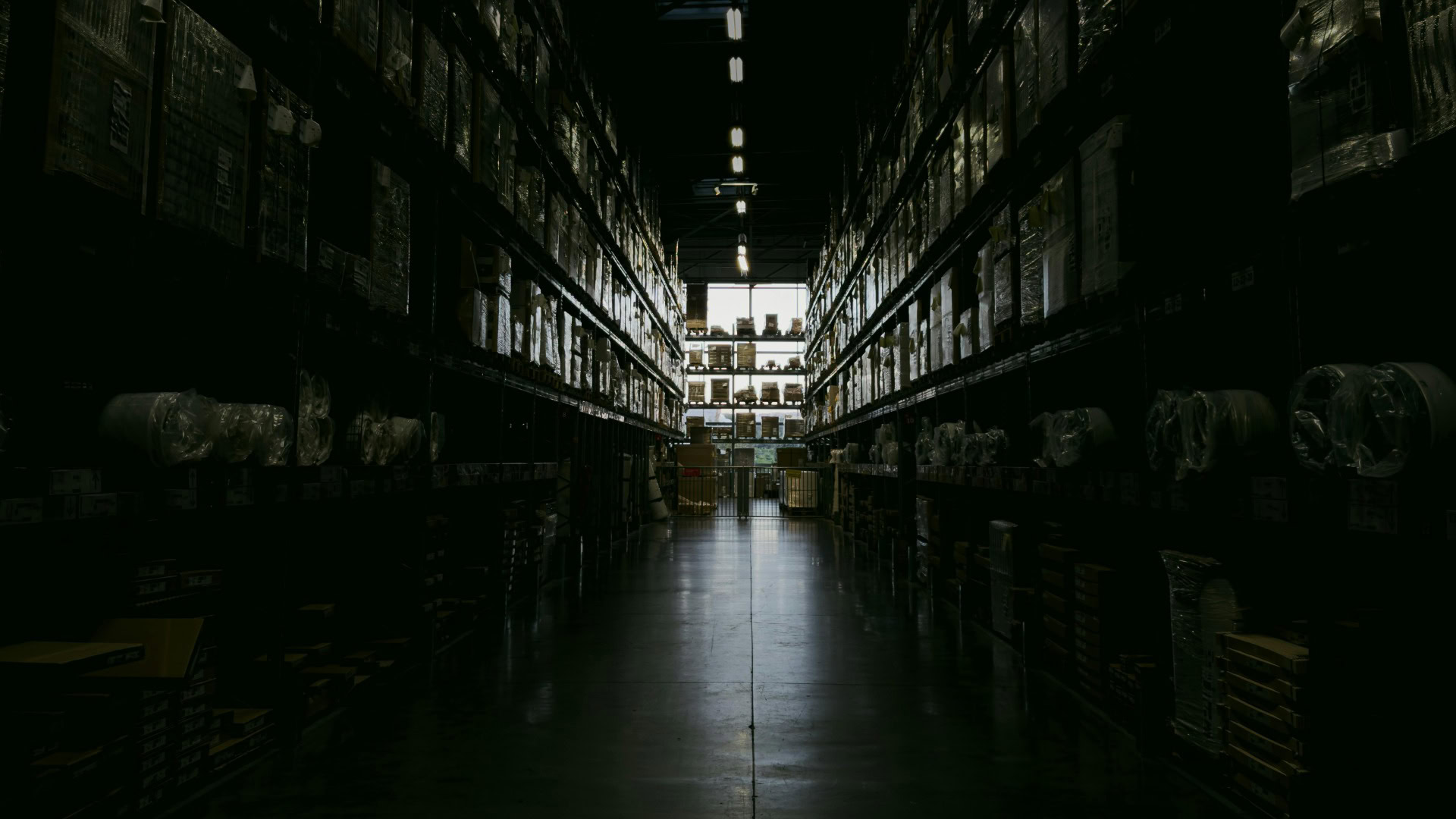 Темний складський прохід із високими стелажами, заповненими коробками та запчастинами, з освітленням у глибині приміщення