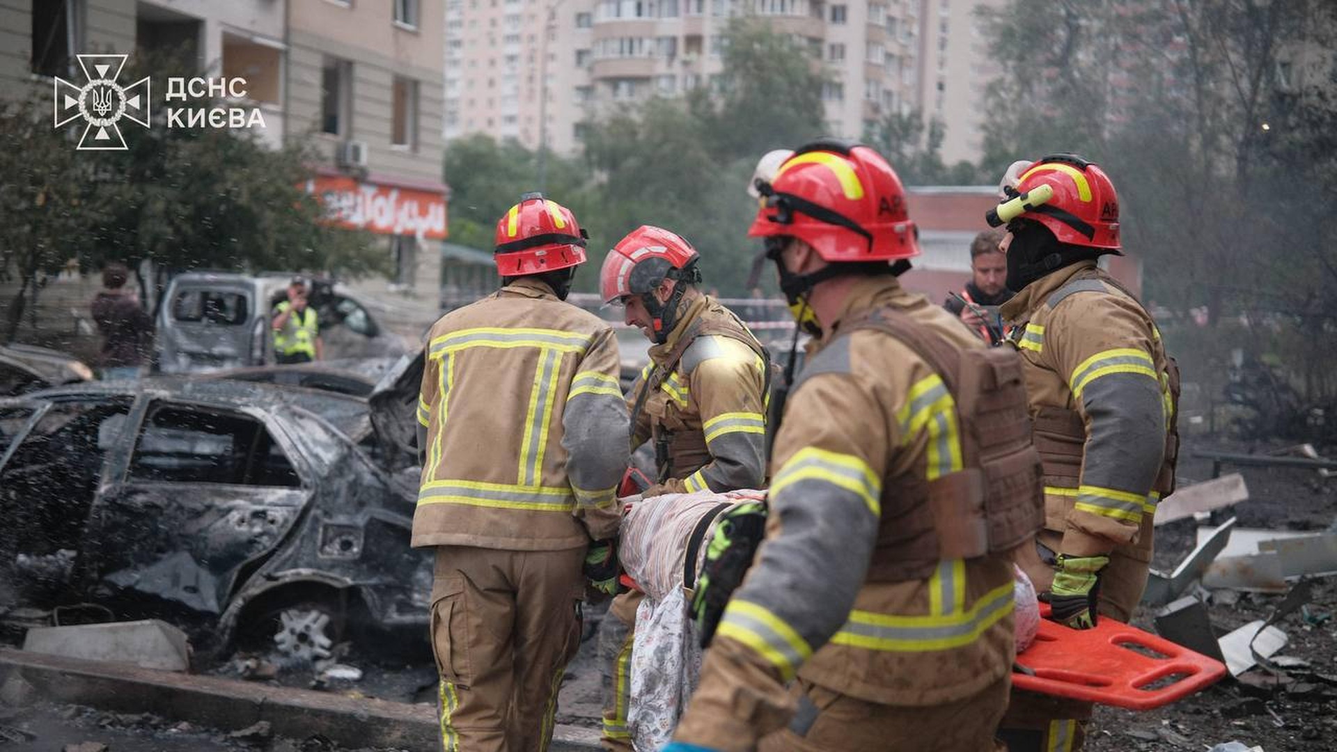 Вишневе: поліція оточила зону ураження — загроза повторної детонації Четверо рятувальників ДСНС несуть постраждалого на ношах повз згорілий автомобіль