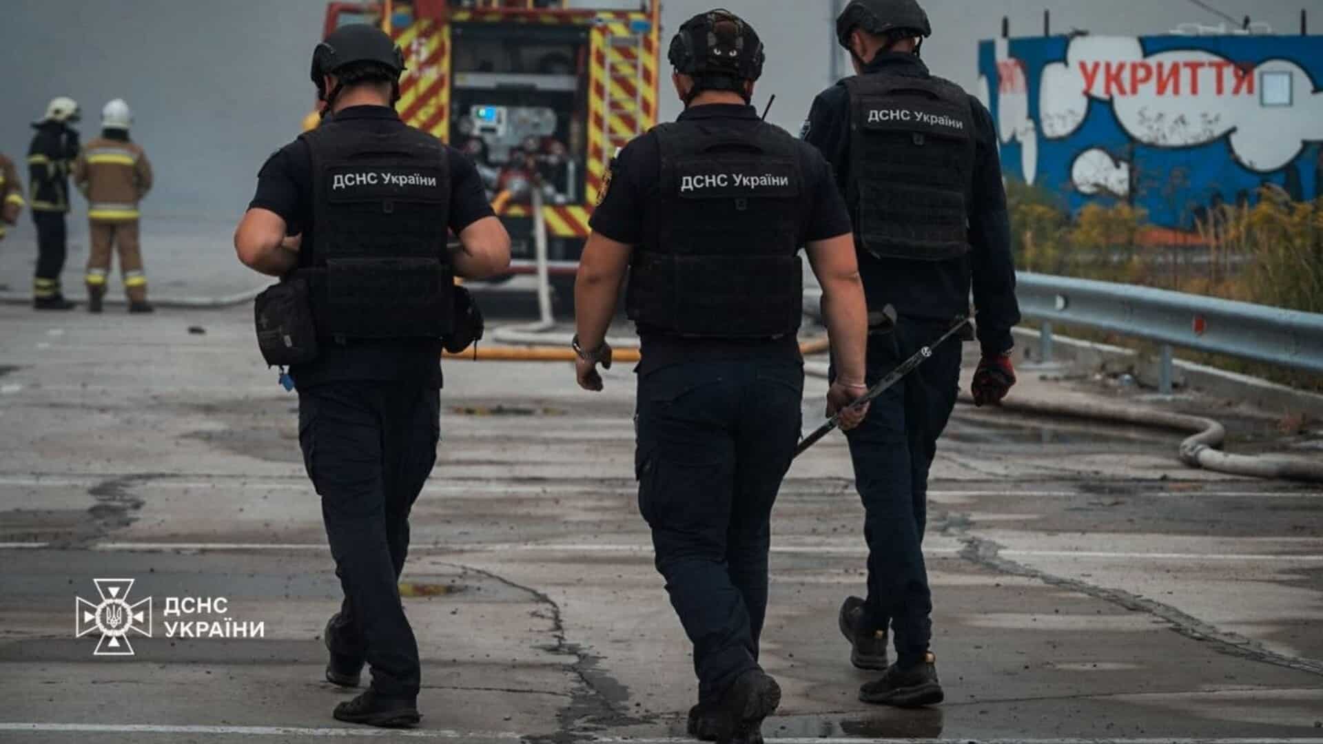 У Борисполі ворожа атака пошкодила дах 9-поверхівки — спалахнула пожежа Троє рятувальників ДСНС України у захисних касках йдуть повз пожежну машину до місця займання, на задньому плані вивіска укриття